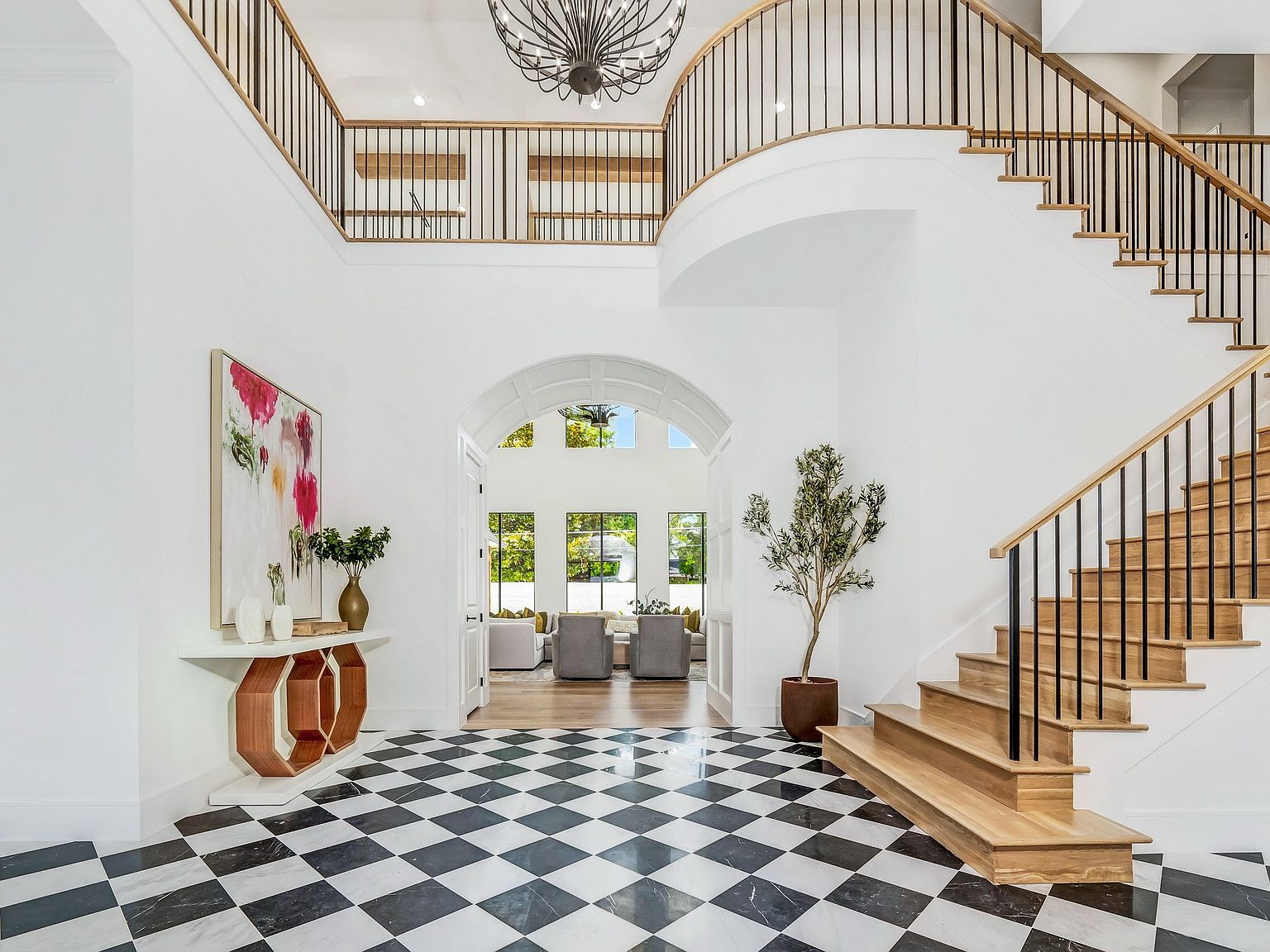 This grand foyer features a striking black and white checkered marble floor, a curved staircase with dark metal railings and light wood steps, and a large arched doorway leading to another room. A modern console table with an abstract painting and a potted olive tree add to the elegance of the space. The high ceilings and abundant natural light create a bright and inviting atmosphere.