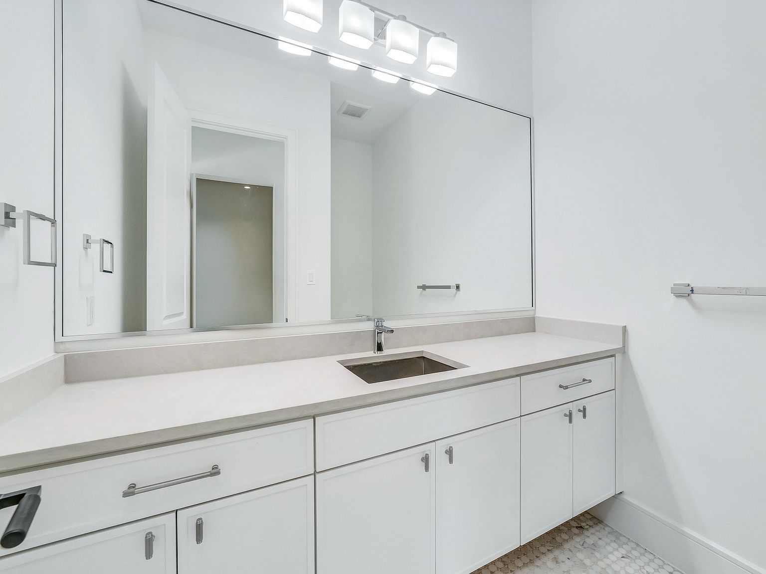 This is a well-lit bathroom featuring a large mirror above a white vanity with a light countertop and a stainless steel sink. The cabinets are white with modern silver hardware. The walls are painted white, and the floor appears to be tiled with small, light-colored tiles.