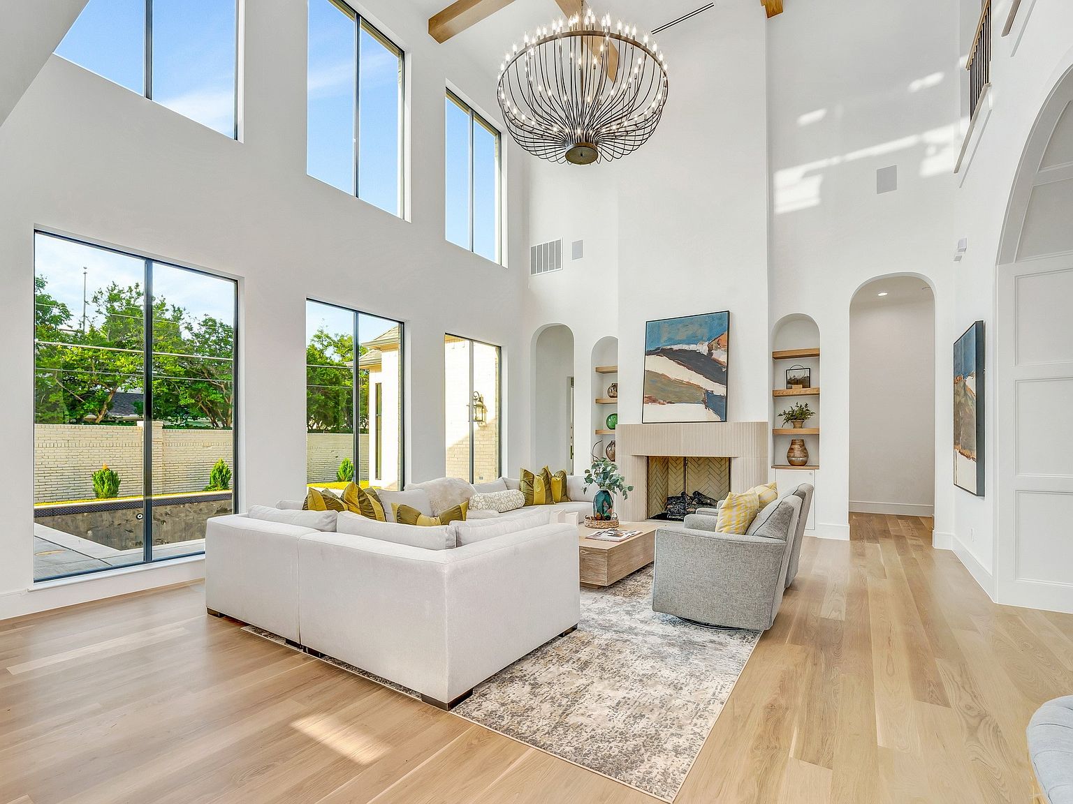 This is a bright and airy living room with soaring ceilings and large windows that flood the space with natural light. The room features a neutral color palette, a large white sofa, a fireplace with a stone surround, and hardwood floors. The overall impression is one of luxury and sophistication.