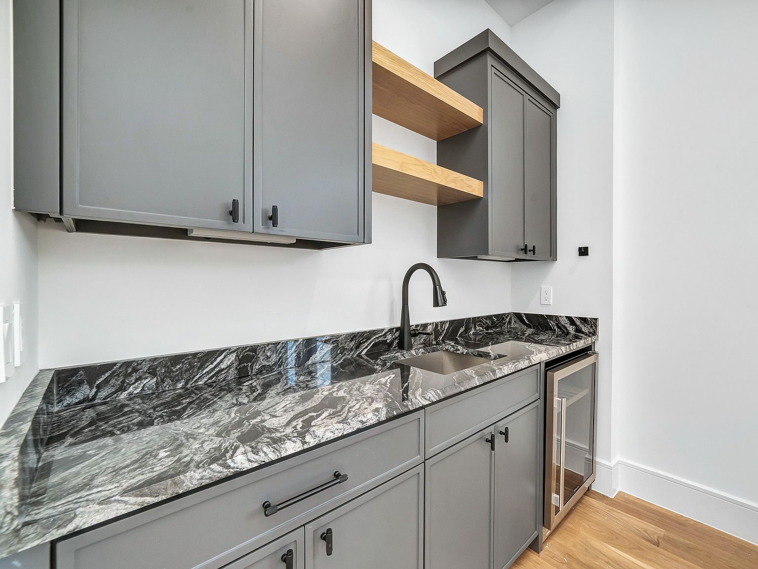 This interior shot showcases a modern kitchen or bar area with gray cabinetry, black hardware, and a striking black and gray marble countertop. A stainless steel sink and a beverage refrigerator are integrated into the design. The space is well-lit, highlighting the clean lines and contemporary aesthetic, perfect for entertaining or everyday use.