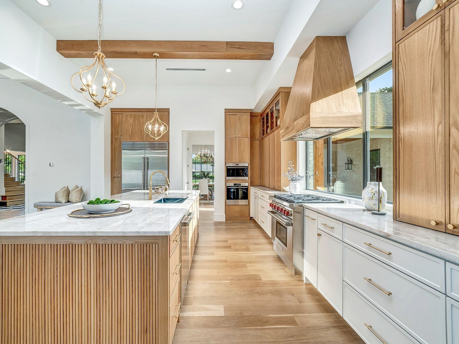This is a bright and spacious kitchen featuring a large island with a marble countertop and wooden base. The kitchen is equipped with stainless steel appliances, custom cabinetry, and a modern range hood. Natural light floods the space through a large window, highlighting the hardwood floors and creating a warm and inviting atmosphere.