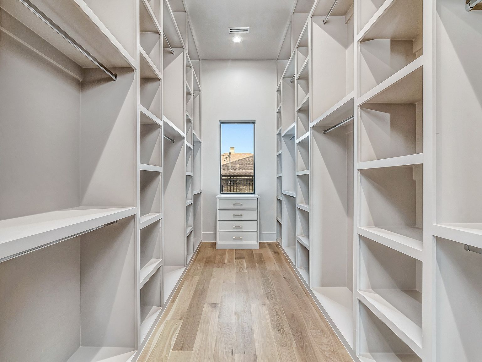 This is a well-organized walk-in closet featuring custom shelving and hanging rods on both sides. A small window at the end of the closet provides natural light, illuminating the light wood flooring and a built-in dresser. The closet is painted in a neutral color, creating a clean and spacious feel.