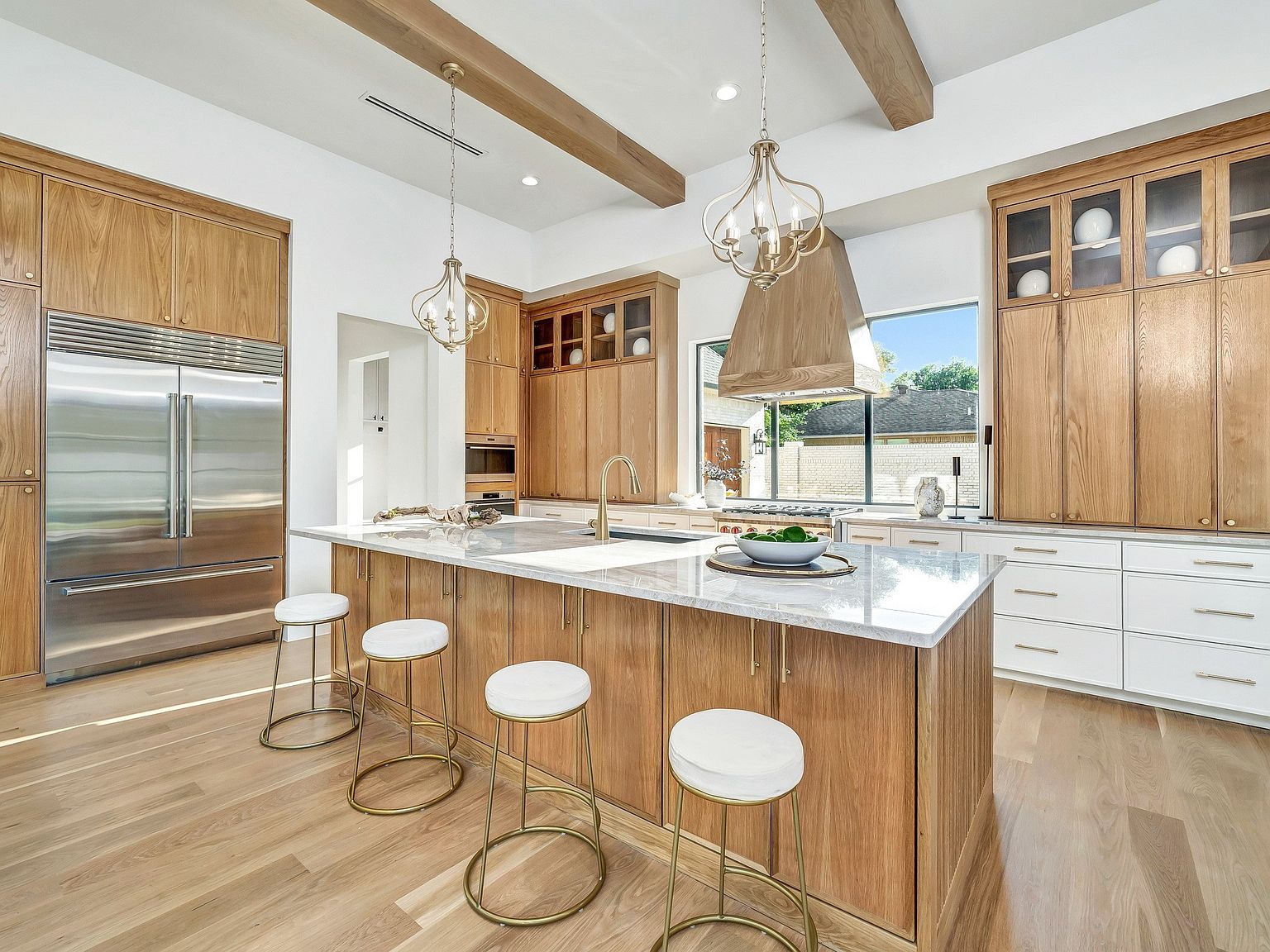 This is a bright and spacious kitchen featuring custom wood cabinetry, a large center island with a marble countertop, and stainless steel appliances. The kitchen is illuminated by pendant lighting and natural light from a large window, creating a warm and inviting atmosphere. The design incorporates modern elements with a touch of rustic charm, making it a desirable space for cooking and entertaining.