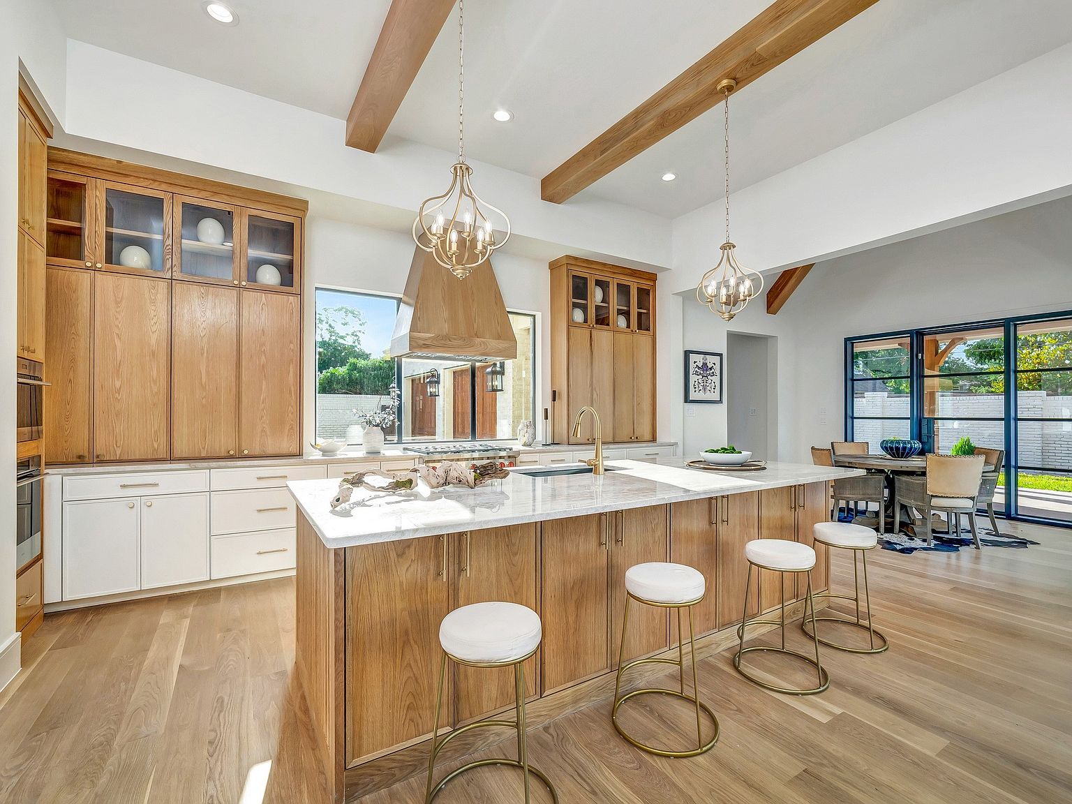 This is a bright and spacious kitchen featuring custom wood cabinetry, a large island with a marble countertop, and modern pendant lighting. The kitchen also includes stainless steel appliances and a view to the outside. The open floor plan seamlessly connects the kitchen to the dining area, creating an inviting space for entertaining.