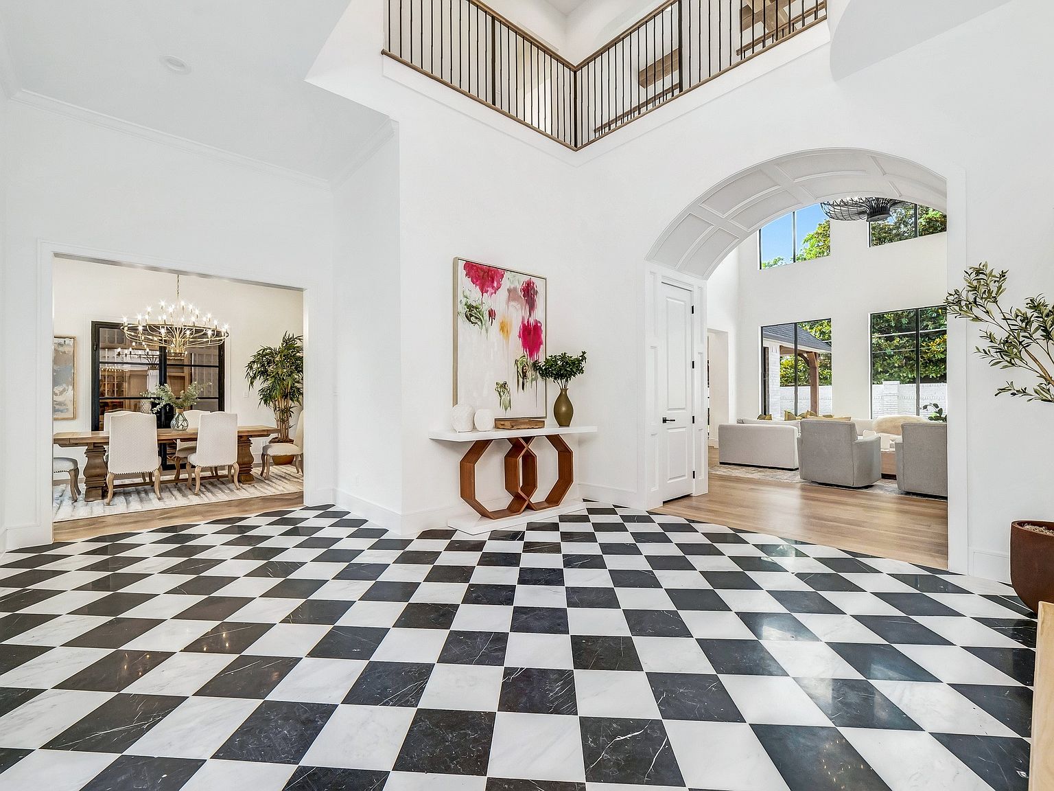 This grand foyer features a striking black and white checkered marble floor, leading into an open-concept living space. A statement art piece hangs above a modern console table, while an arched doorway provides a glimpse into a bright living room. The high ceilings and wrought iron balcony add to the luxurious feel of the home.