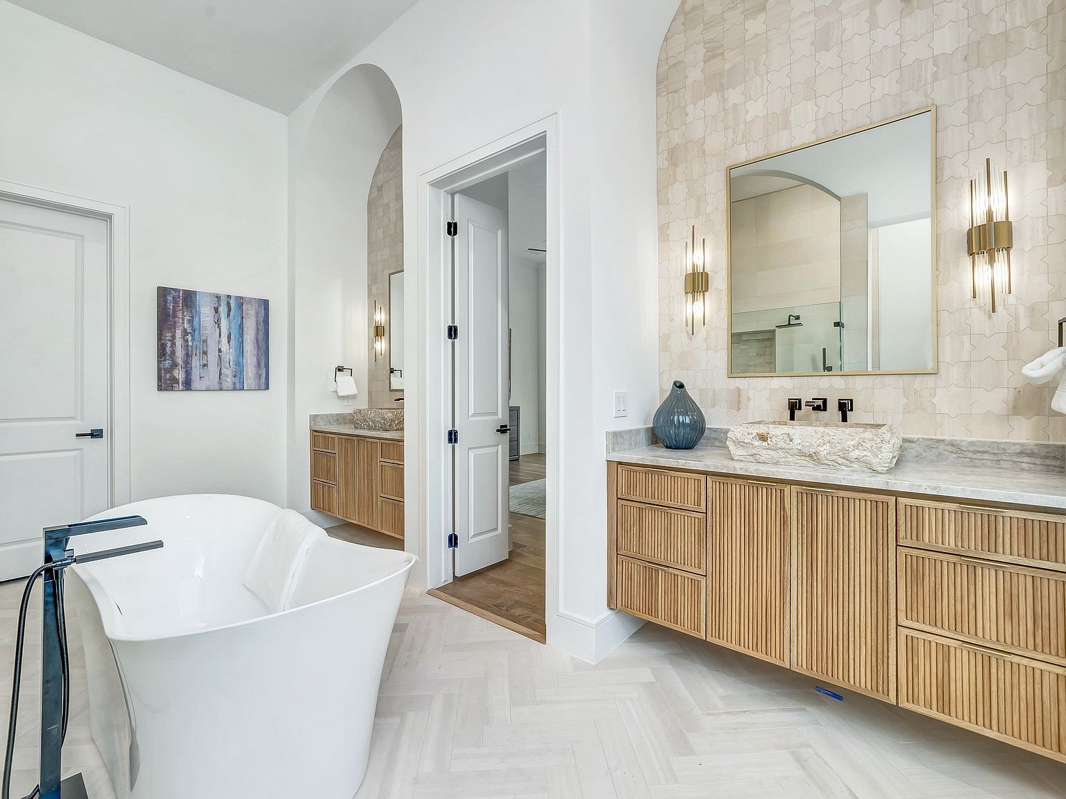 This is a luxurious primary bathroom featuring a freestanding white bathtub, herringbone-patterned light wood flooring, and dual vanities with light wood cabinetry and stone countertops. The walls are a mix of white and patterned tile, and the space is illuminated by modern sconces, creating a bright and elegant atmosphere. The perspective is from the doorway, showcasing the depth and design of the bathroom.