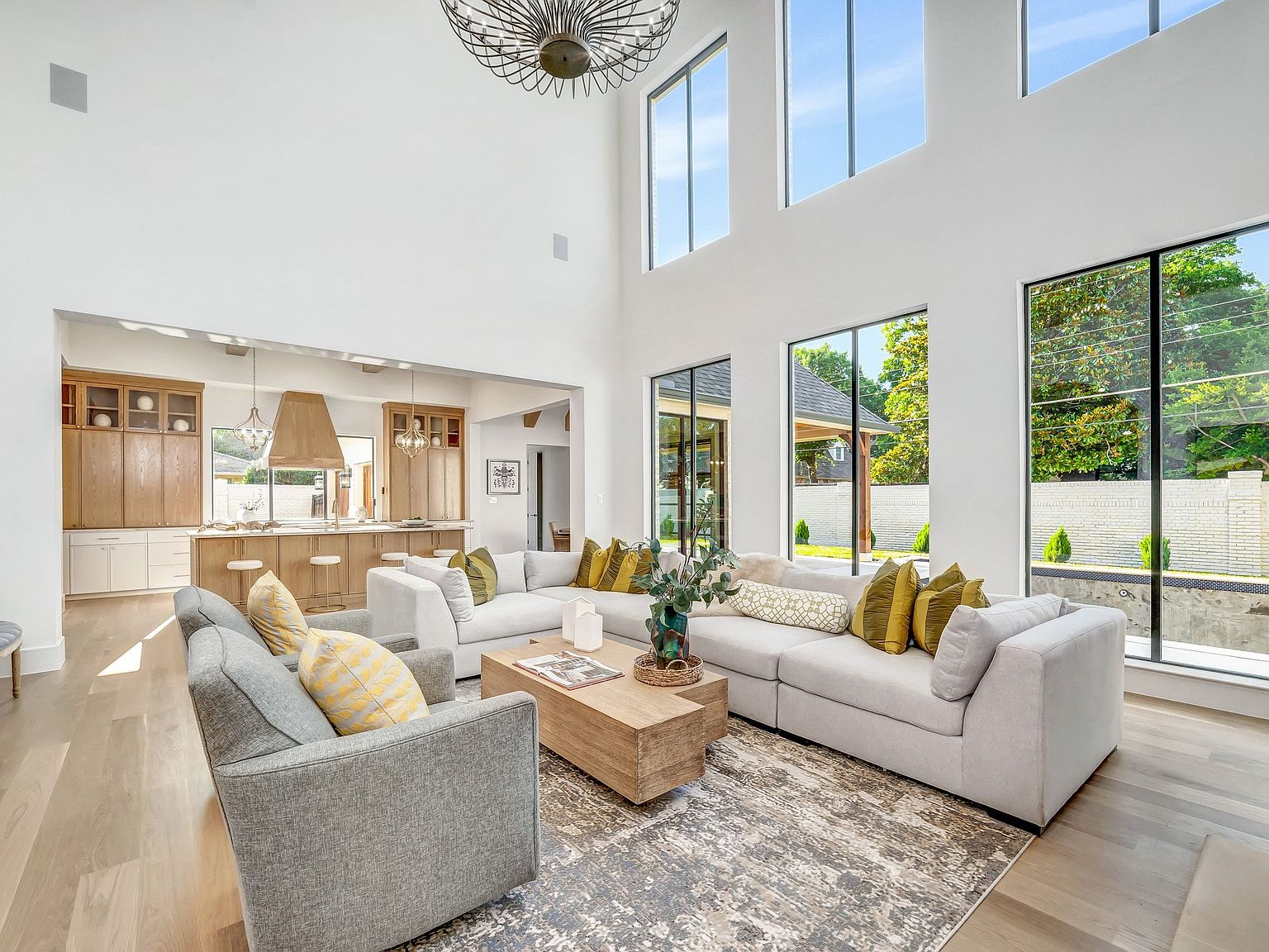 This is an interior shot of a modern living room featuring a large sectional sofa with decorative pillows, a wooden coffee table, and a gray armchair. The room has high ceilings with tall windows that provide ample natural light and views of the exterior landscape. The open-concept design seamlessly connects the living room to the kitchen, creating a spacious and inviting atmosphere.