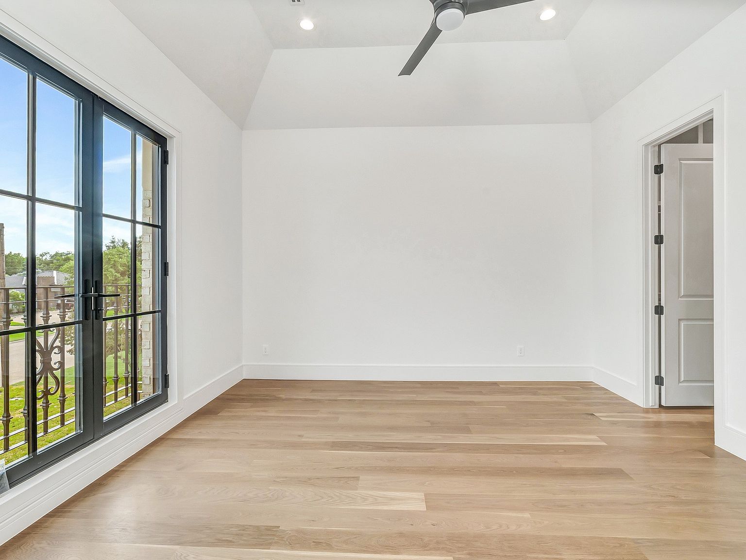 This is a bright and airy bedroom featuring hardwood floors, white walls, and a large window with black frames that offers a view of the outdoors. The room has a minimalist aesthetic with a ceiling fan and a doorway leading to another room. The space feels clean, modern, and inviting.