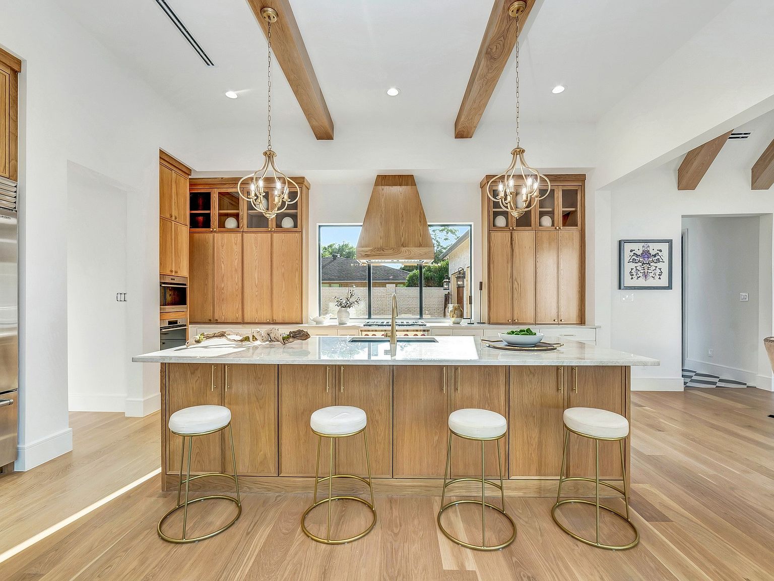 This is a bright and spacious kitchen featuring light wood cabinetry, a large island with white countertops, and four bar stools with gold bases and white seats. The kitchen is illuminated by pendant lighting and natural light from a window above the stove. The overall style is modern and inviting, with a focus on natural materials and clean lines.