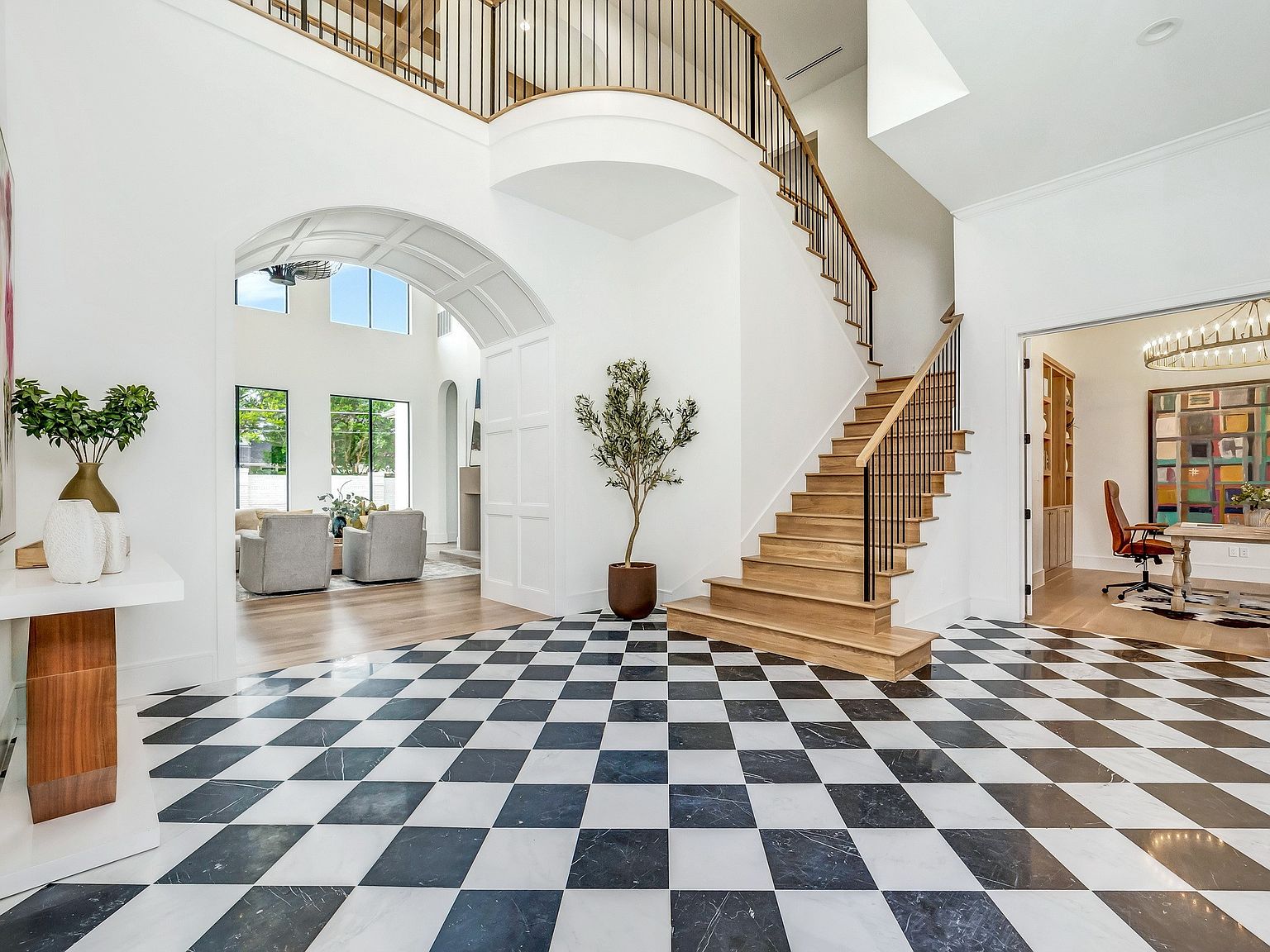This grand foyer features a striking black and white checkered marble floor, leading to a staircase with wooden steps and black iron railings. The space is bright and airy, with high ceilings and an arched entryway leading to a living area. A potted olive tree adds a touch of greenery, and a glimpse into an office space can be seen to the right.