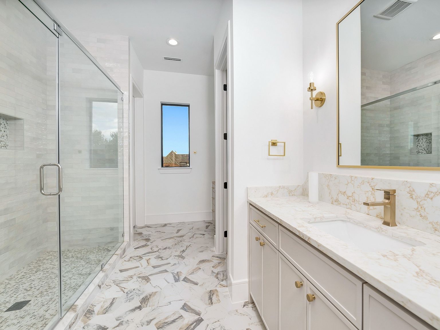 This is a bright and modern primary bathroom featuring a glass-enclosed shower, a vanity with a marble countertop, and gold fixtures. The flooring is a stylish marble tile, and a window provides natural light. The overall impression is clean, luxurious, and well-designed.