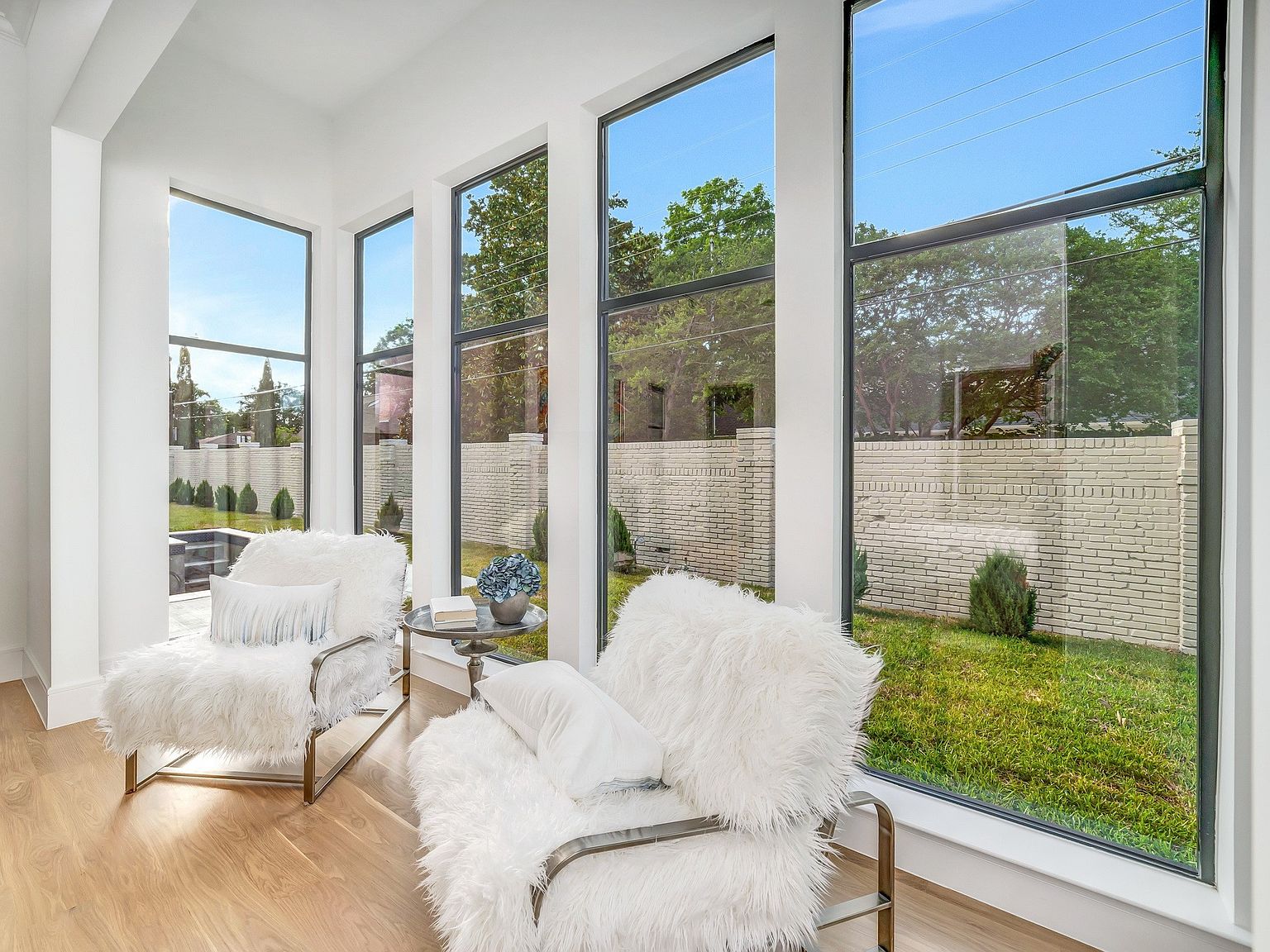 This interior shot showcases a bright and airy living room with large windows offering views of the exterior landscape. Two plush, white faux fur chairs with chrome frames are positioned to enjoy the natural light and scenery. A small side table with a floral arrangement adds a touch of elegance to the space, which is characterized by its modern design and hardwood flooring.