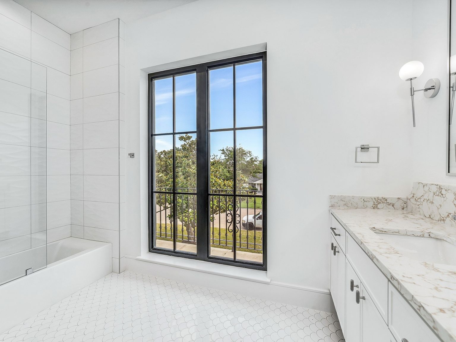 This is a bright and airy primary bathroom featuring a large window with black frames offering a view of the outdoors. The bathroom includes a white vanity with a marble countertop, white cabinetry, and modern fixtures. The floor is tiled with a decorative pattern, and the walls are painted white, creating a clean and luxurious atmosphere.