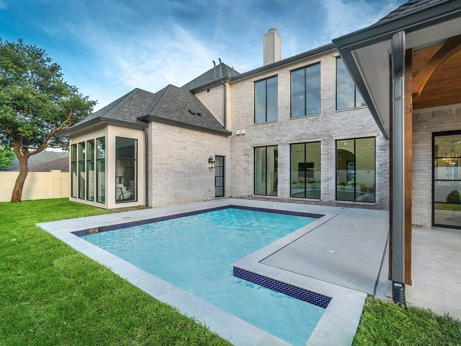 This image showcases the rear exterior of a luxurious home, highlighting a modern swimming pool with a tiled edge and a concrete patio area. The house features light-colored brick, large windows, and a dark roof, creating a sophisticated and inviting outdoor living space. Lush green grass surrounds the pool area, adding to the overall appeal.