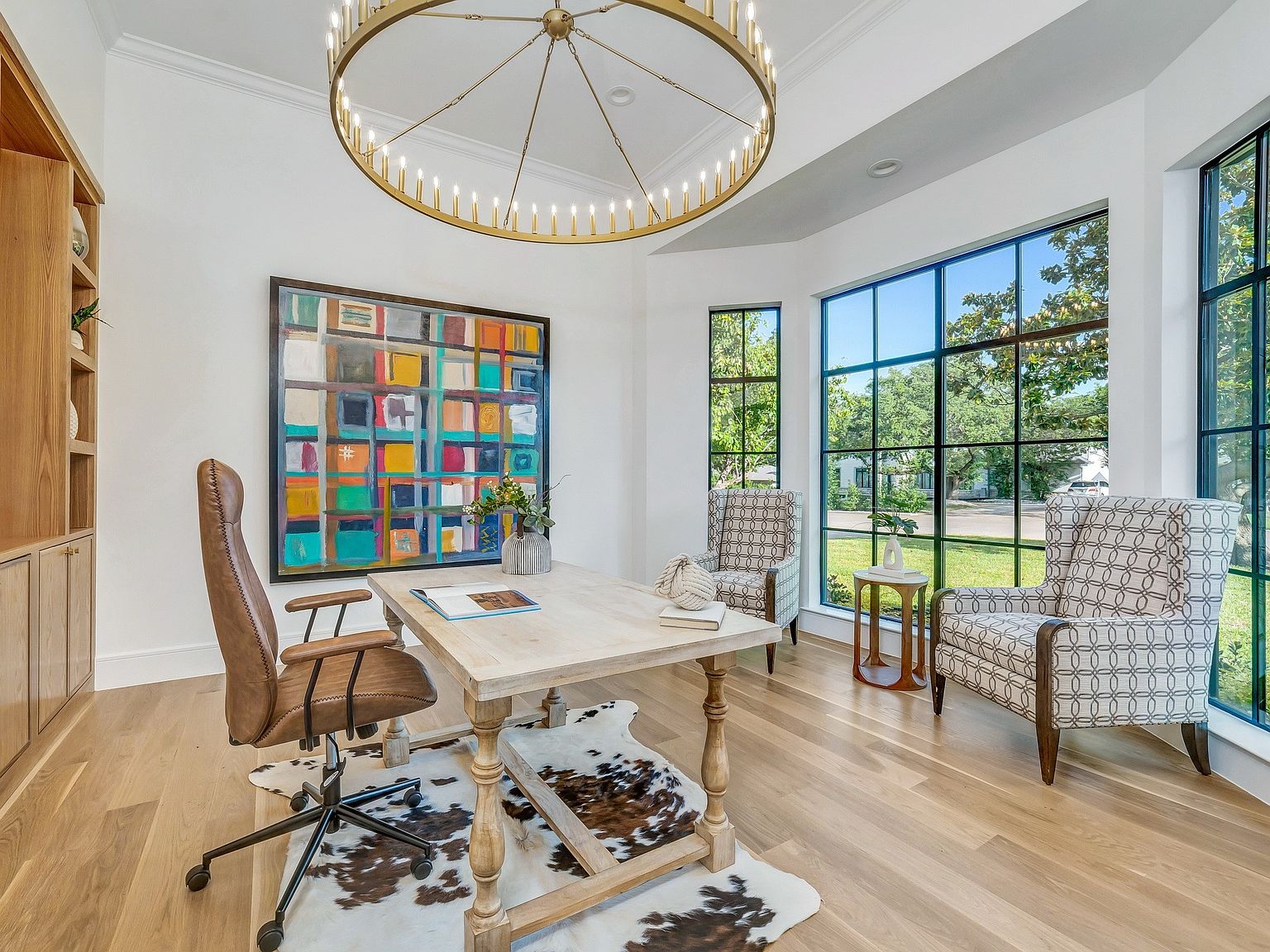 This is an interior shot of a well-lit home office. The room features a large, colorful abstract painting, a wooden desk with a cowhide rug underneath, and two patterned armchairs near a large window offering a view of the outdoors. A striking chandelier hangs above, adding a touch of elegance to the workspace.