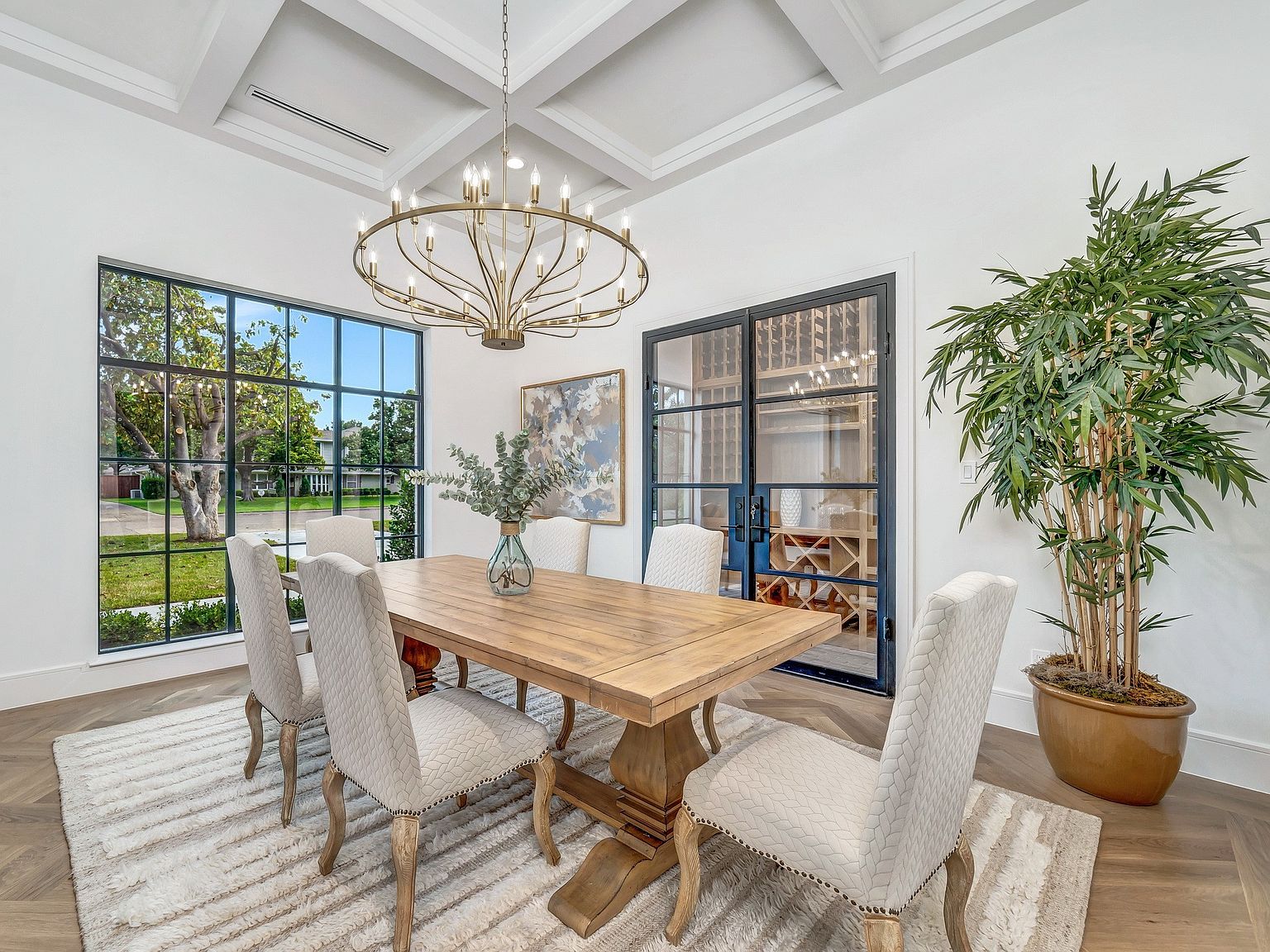 This is an interior shot of a dining room featuring a large wooden table with six upholstered chairs. A modern chandelier hangs above the table, and a large window provides natural light and a view of the outdoors. A wine cellar is visible through a glass-paned door, and a large potted plant adds a touch of greenery to the space, creating an elegant and inviting atmosphere.