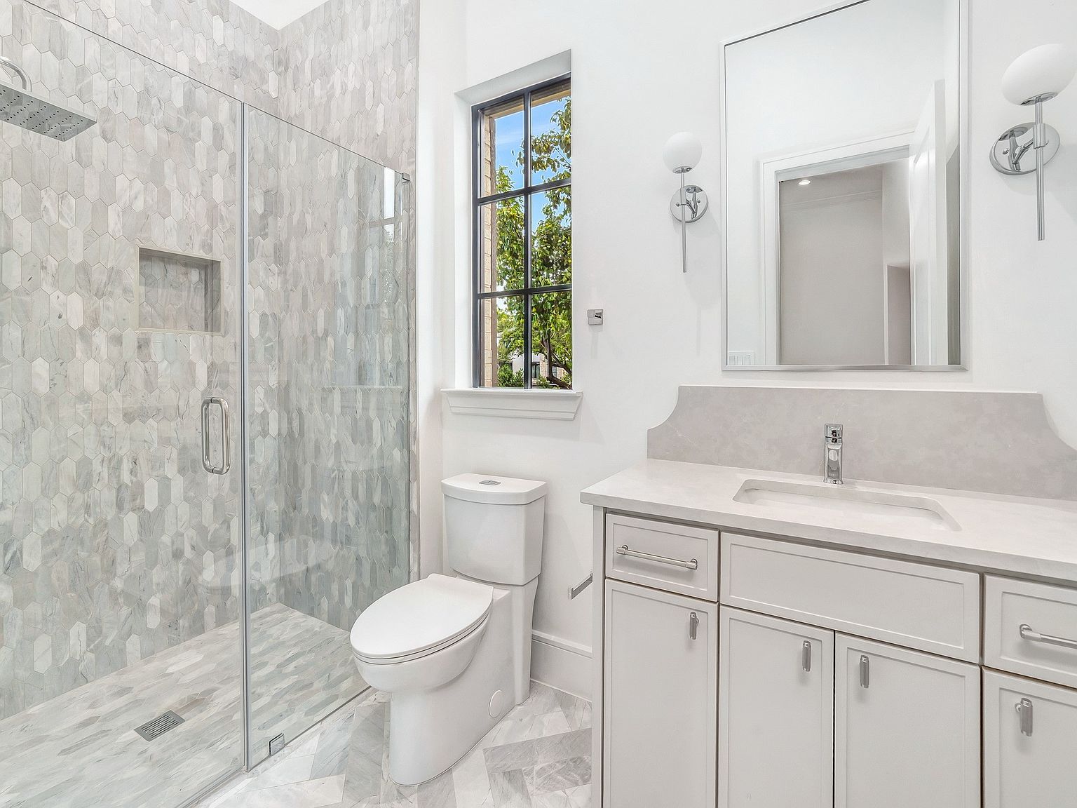 This is a well-lit bathroom featuring a glass-enclosed shower with marble-like tiling, a toilet, and a vanity with white cabinetry and a light-colored countertop. The walls are painted white, and there is a window above the toilet, providing natural light. The overall impression is clean, modern, and luxurious.