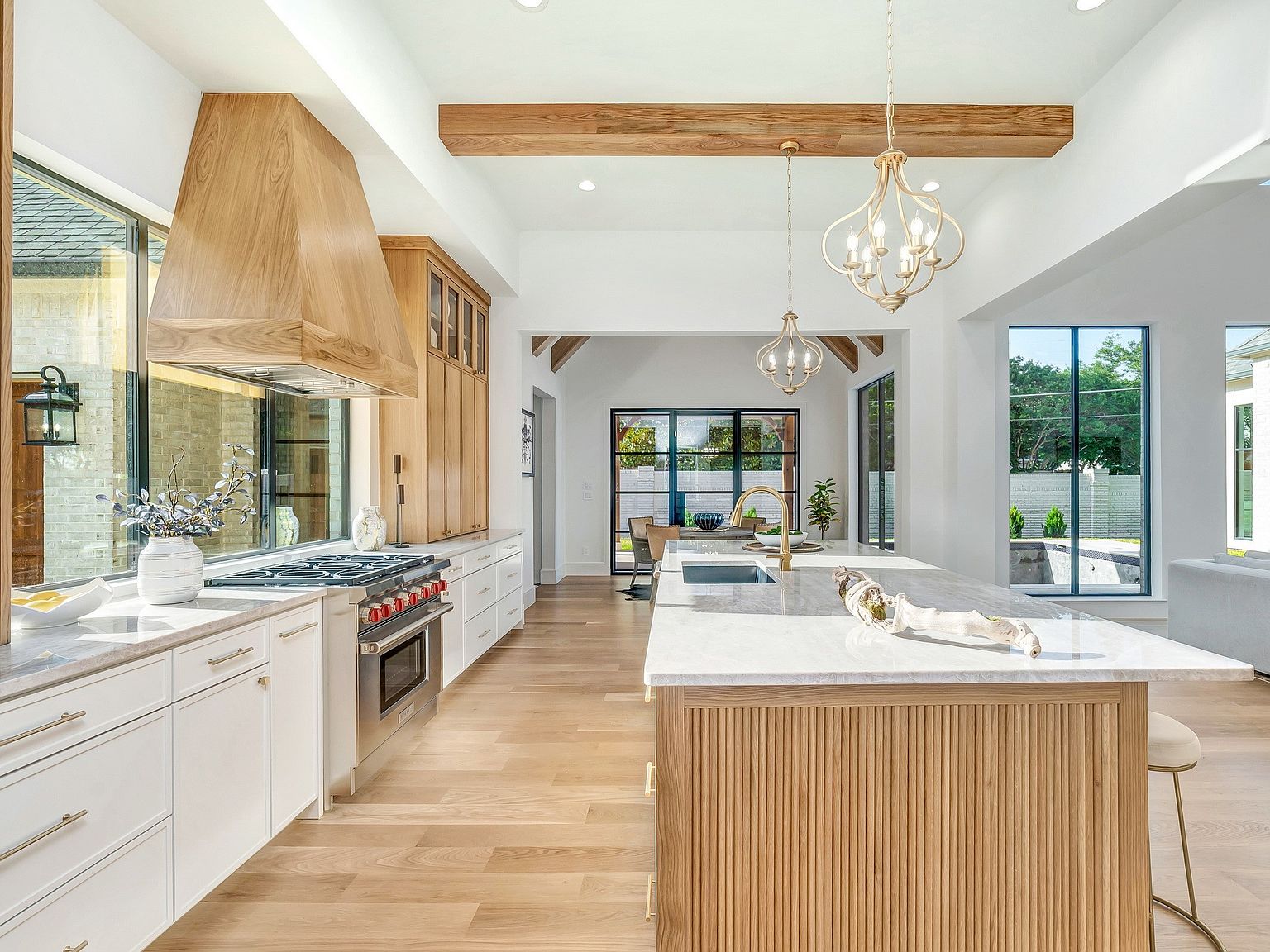 This is a bright and spacious kitchen featuring a large island with a marble countertop and wooden base. The kitchen is equipped with stainless steel appliances, a custom wooden range hood, and white cabinetry. Natural light floods the space through large windows, highlighting the hardwood floors and creating a warm and inviting atmosphere.