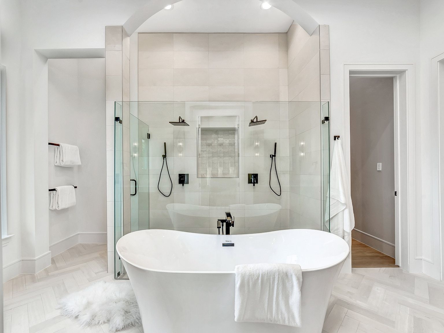 This is a luxurious primary bathroom featuring a freestanding white bathtub as the focal point, complemented by a glass-enclosed walk-in shower with dual shower heads and sleek black fixtures. The space is bright and airy, with light-colored tile flooring laid in a herringbone pattern and neutral wall tones, creating a spa-like atmosphere. The perspective is from the entrance of the bathroom, showcasing the tub and shower area.