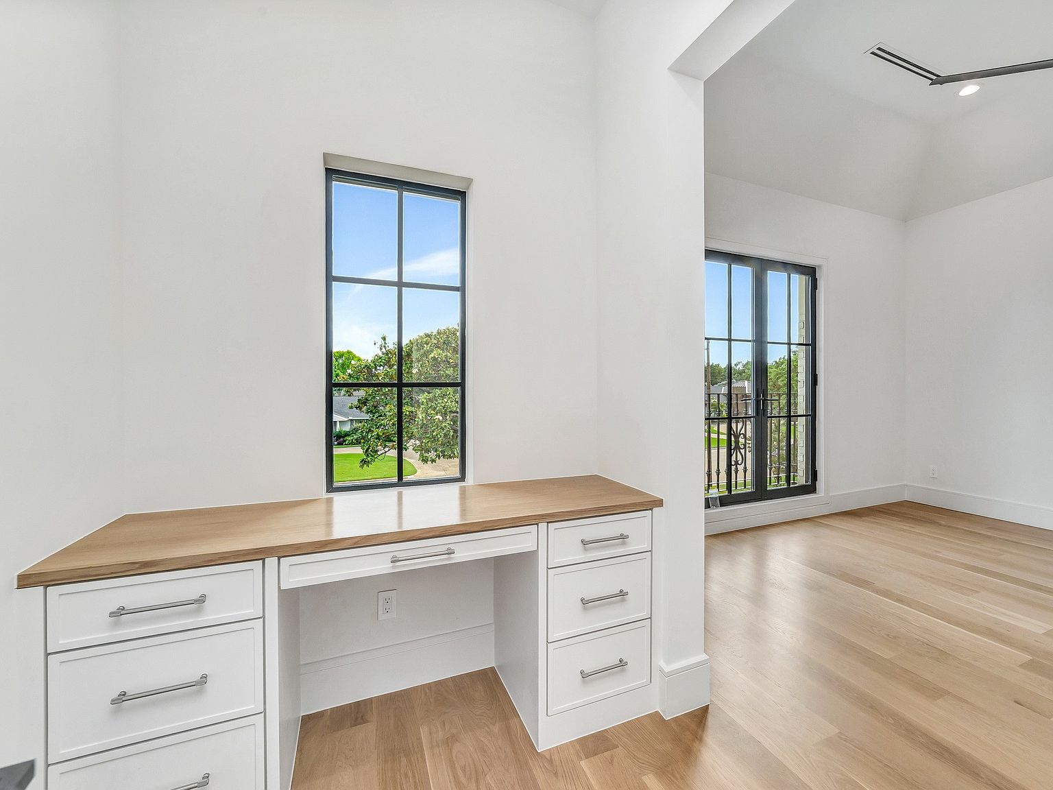 This interior shot showcases a bright and modern office space. A built-in desk with a light wood countertop and white cabinetry sits beneath a window, offering natural light. The room features hardwood flooring and white walls, creating a clean and airy atmosphere, with a glimpse into an adjacent room through a wide doorway.