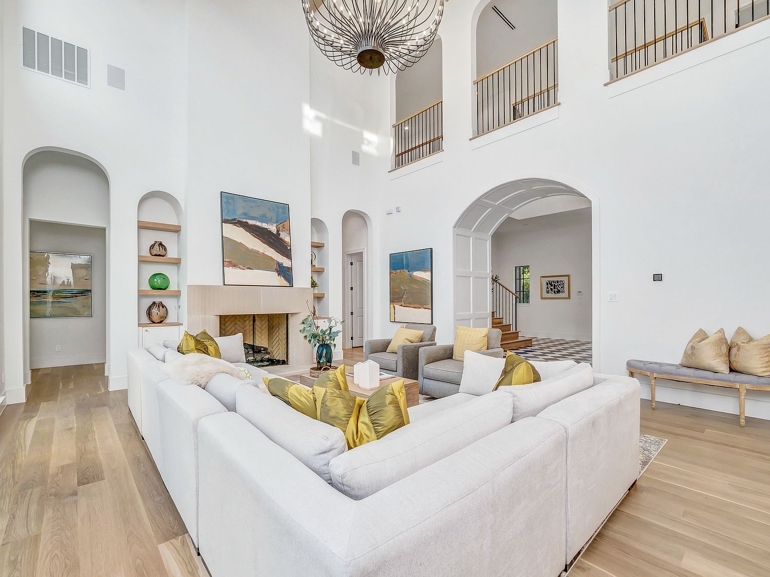 This is a spacious living room featuring a large white sectional sofa with yellow pillows, a stone fireplace with artwork above, and built-in shelving. The room has high ceilings with a decorative chandelier and an open floor plan leading to other areas of the house. The style is modern and luxurious, creating a bright and inviting atmosphere.