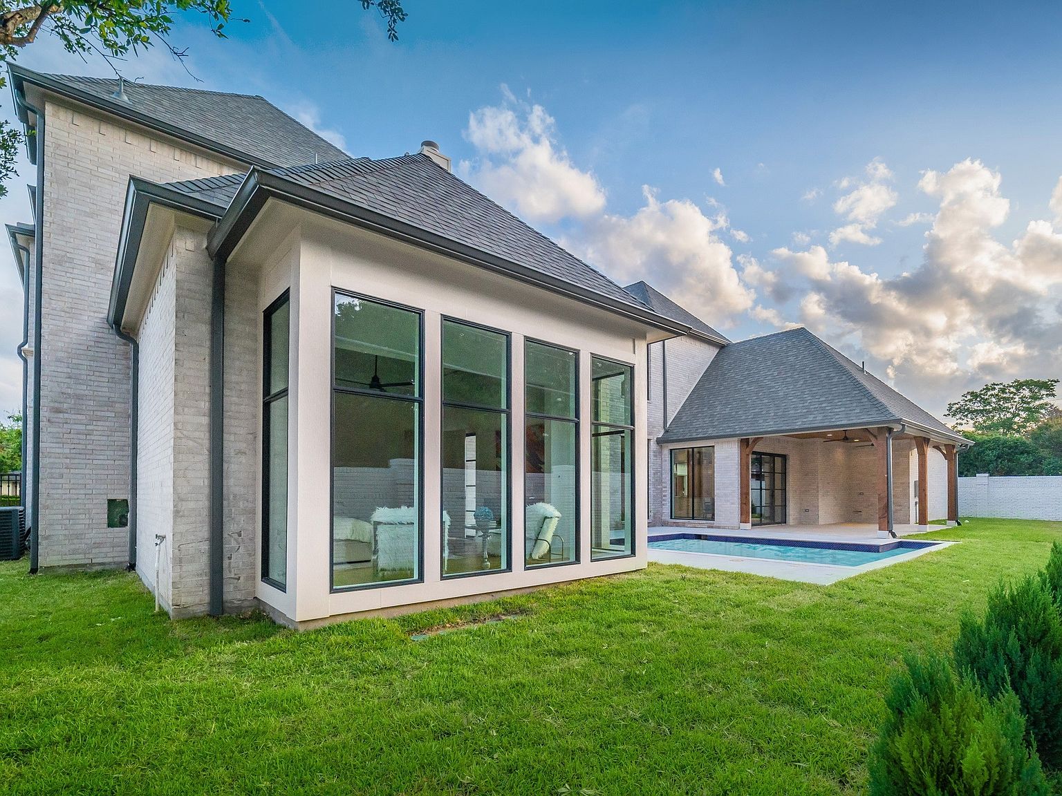 This image showcases the rear exterior of a modern home, featuring a light brick facade, large windows, and a dark gray roof. A pool and an outdoor living area with a covered patio are visible, suggesting a focus on outdoor entertainment and relaxation. The well-maintained lawn and landscaping add to the property's appeal.