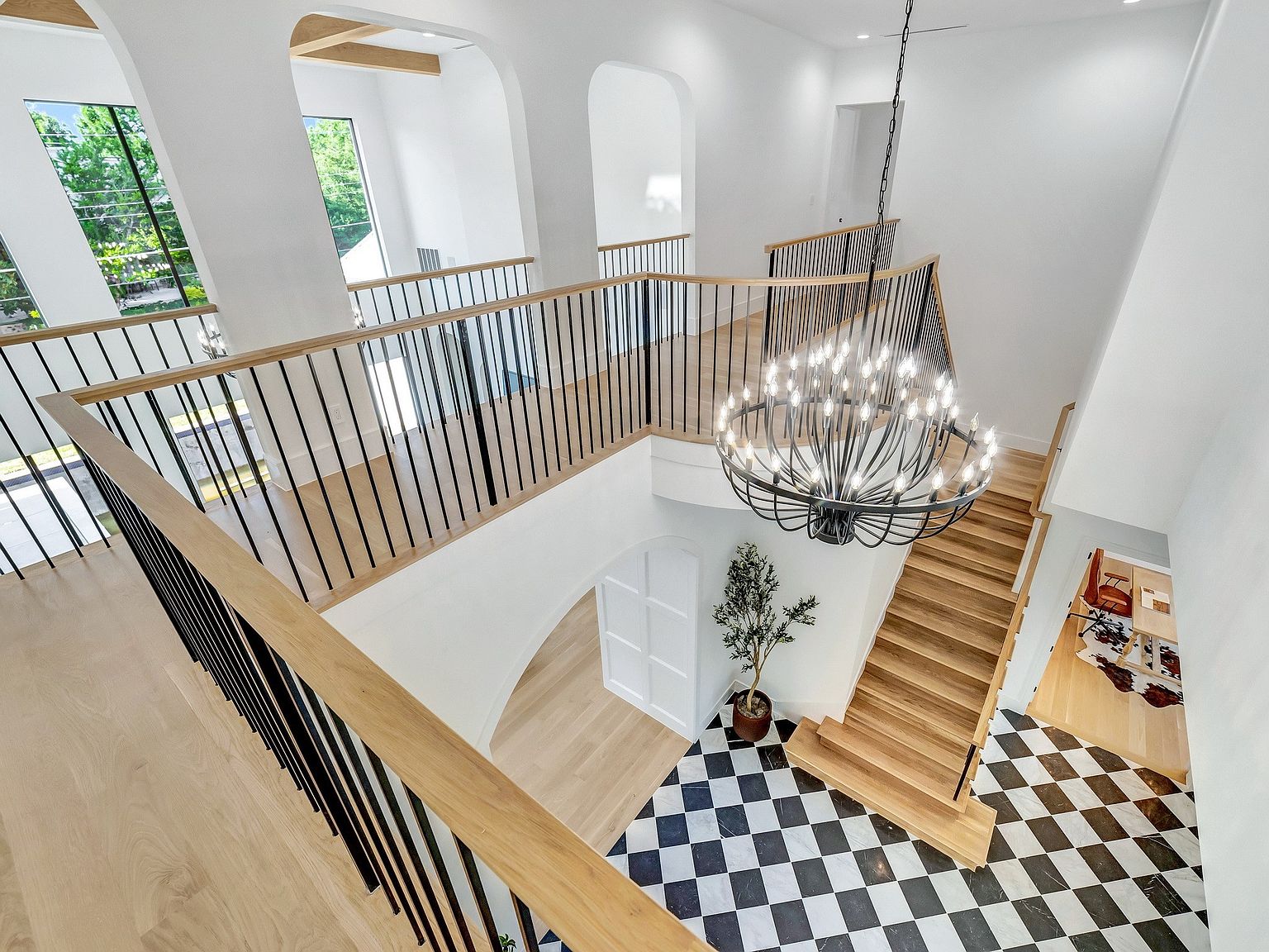 This is an interior shot of a grand foyer featuring a staircase, a large chandelier, and a black and white checkered tile floor. The staircase has wooden steps and leads to a second-floor landing with black metal railings and wooden handrails. The walls are painted white, and the space is well-lit, creating a bright and inviting atmosphere.