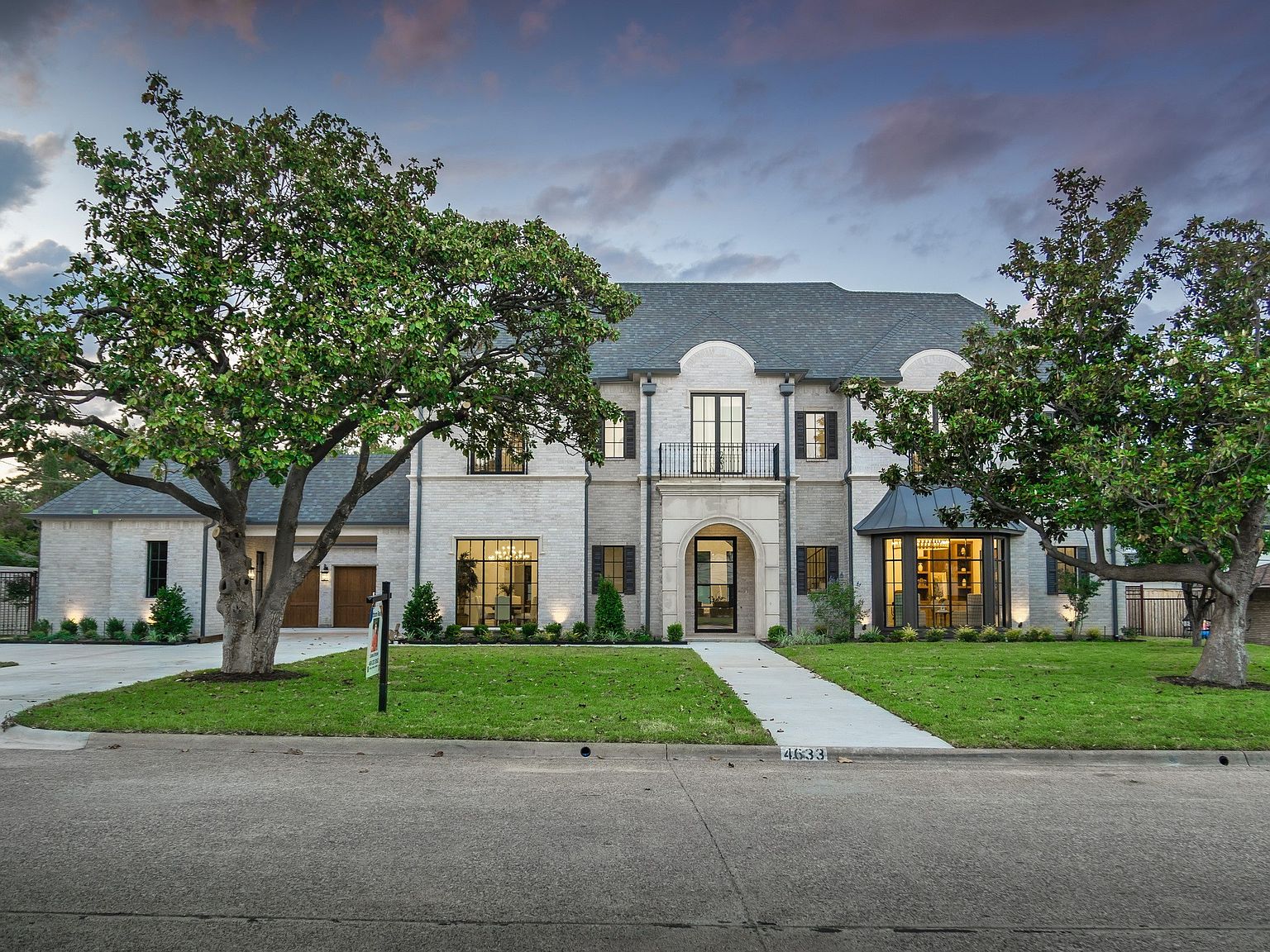 This is a front exterior view of a large, luxurious two-story home. The house features a light-colored brick facade, a dark gray roof, and well-manicured landscaping. Large trees frame the house, adding to its curb appeal and creating a sense of established elegance.