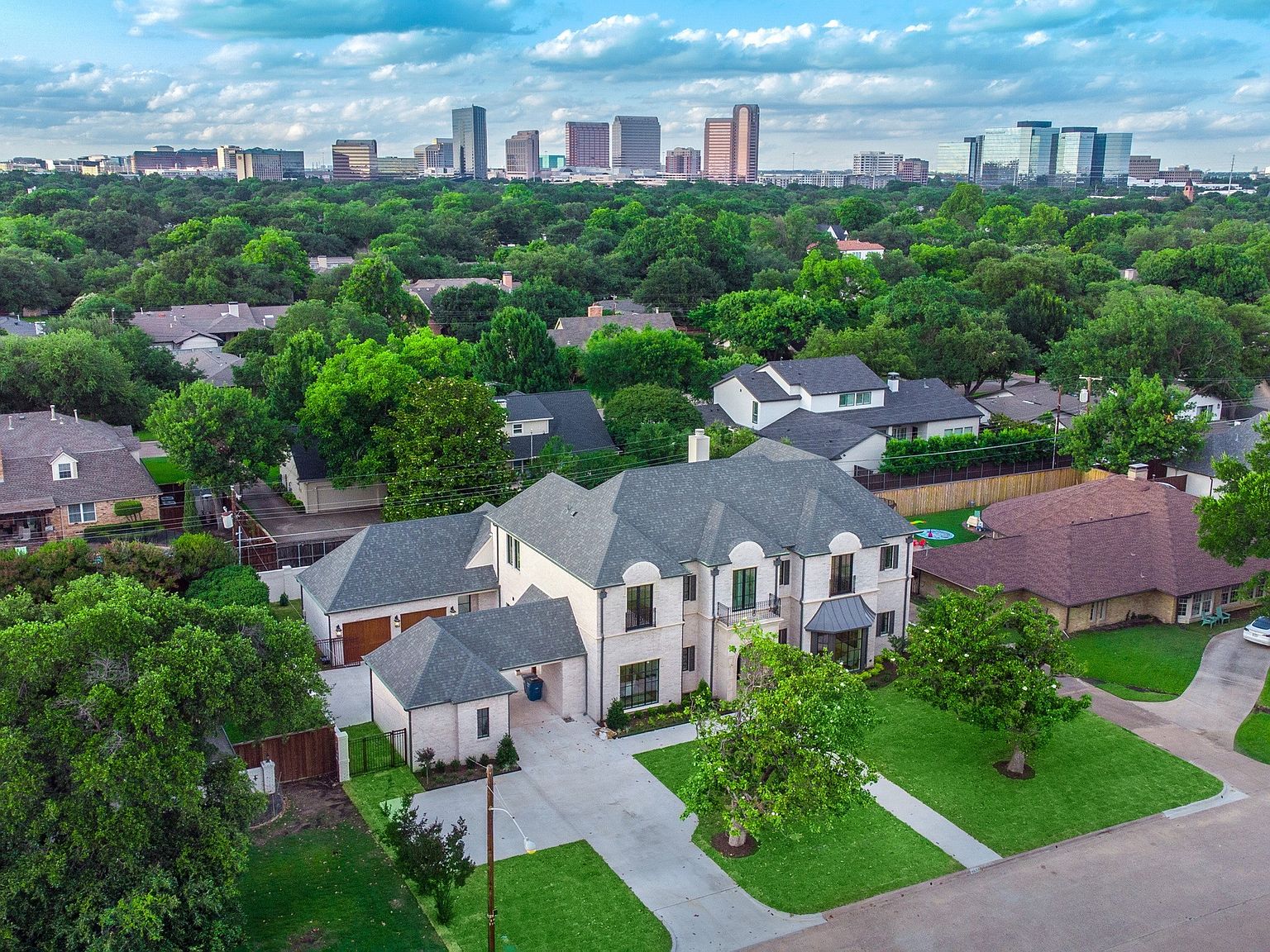 This aerial view showcases a large, luxurious home with a gray roof, light-colored brick exterior, and well-manicured lawn. The property includes a detached garage and is surrounded by mature trees, offering privacy. In the background, a city skyline adds a sense of urban proximity.