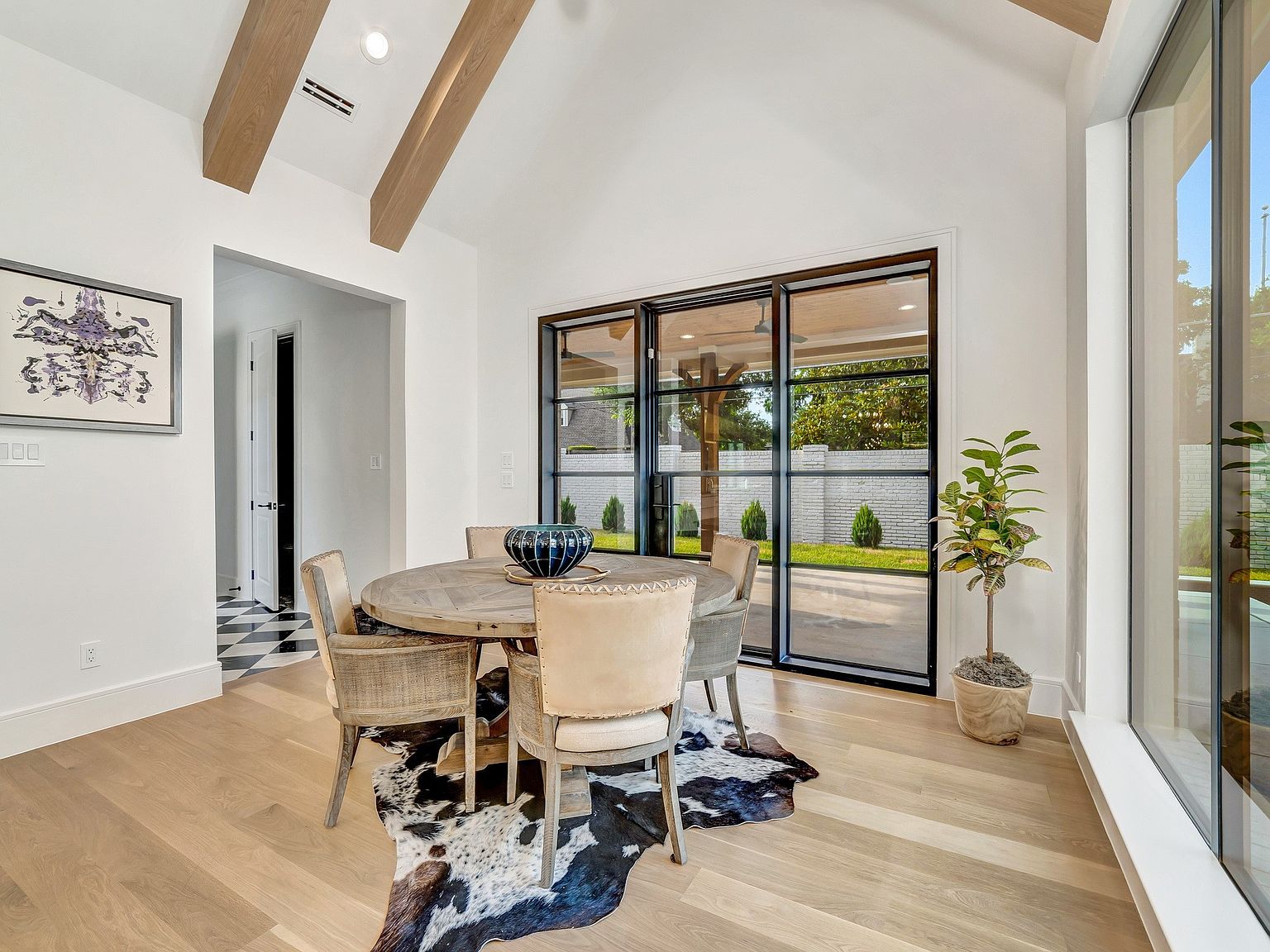 This dining room features a round wooden table surrounded by woven chairs, set upon a cowhide rug. Natural light floods the space through large black-framed windows, highlighting the light wood flooring and white walls. Exposed wooden beams add architectural interest to the high ceiling, creating a bright and inviting atmosphere.