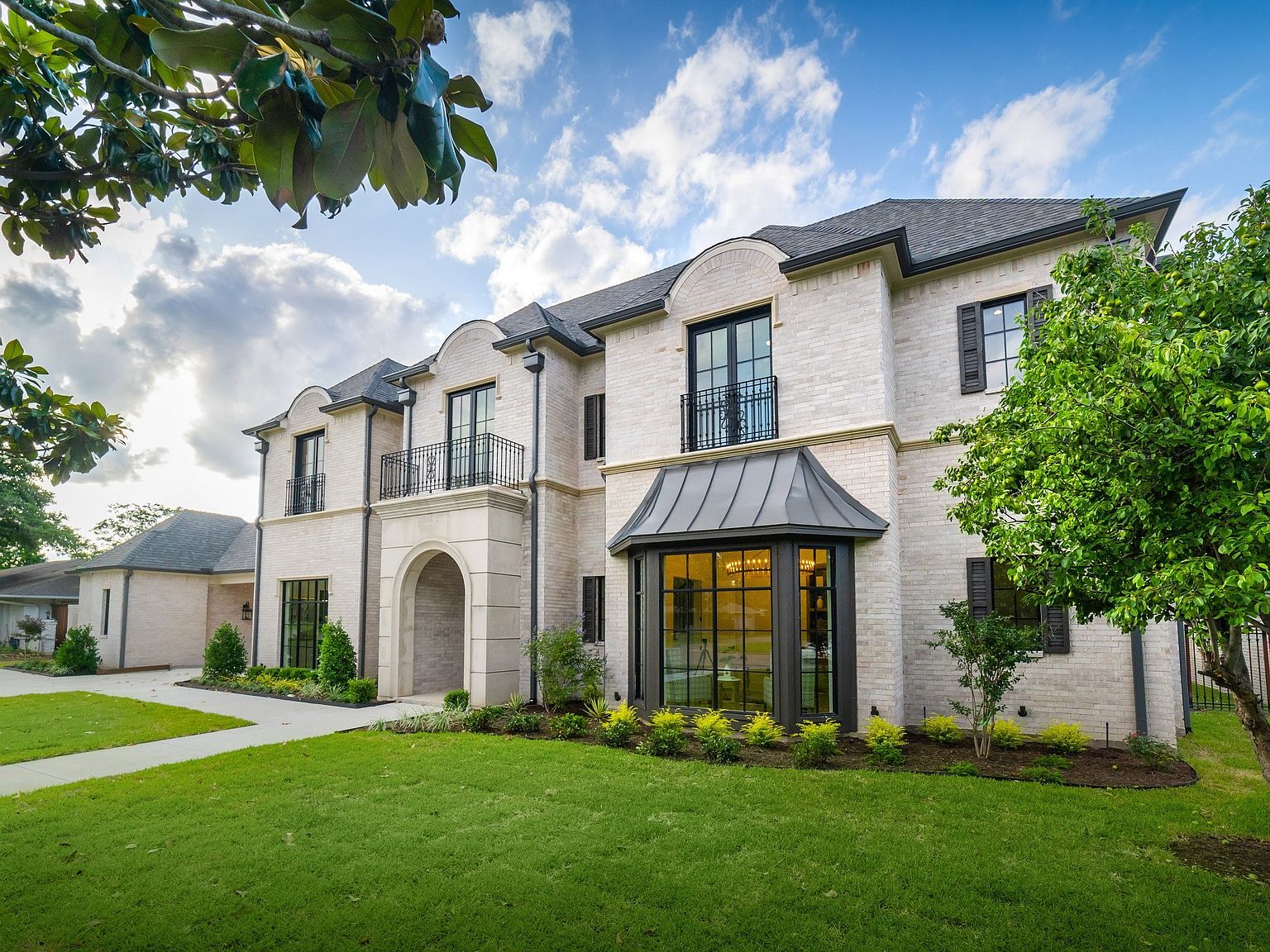 This is a front exterior view of a large, luxurious two-story house with a light-colored brick facade. The house features multiple windows with black frames, some with balconies, and a prominent arched entryway. The manicured lawn and landscaping enhance the property's curb appeal, creating an impression of elegance and sophistication.