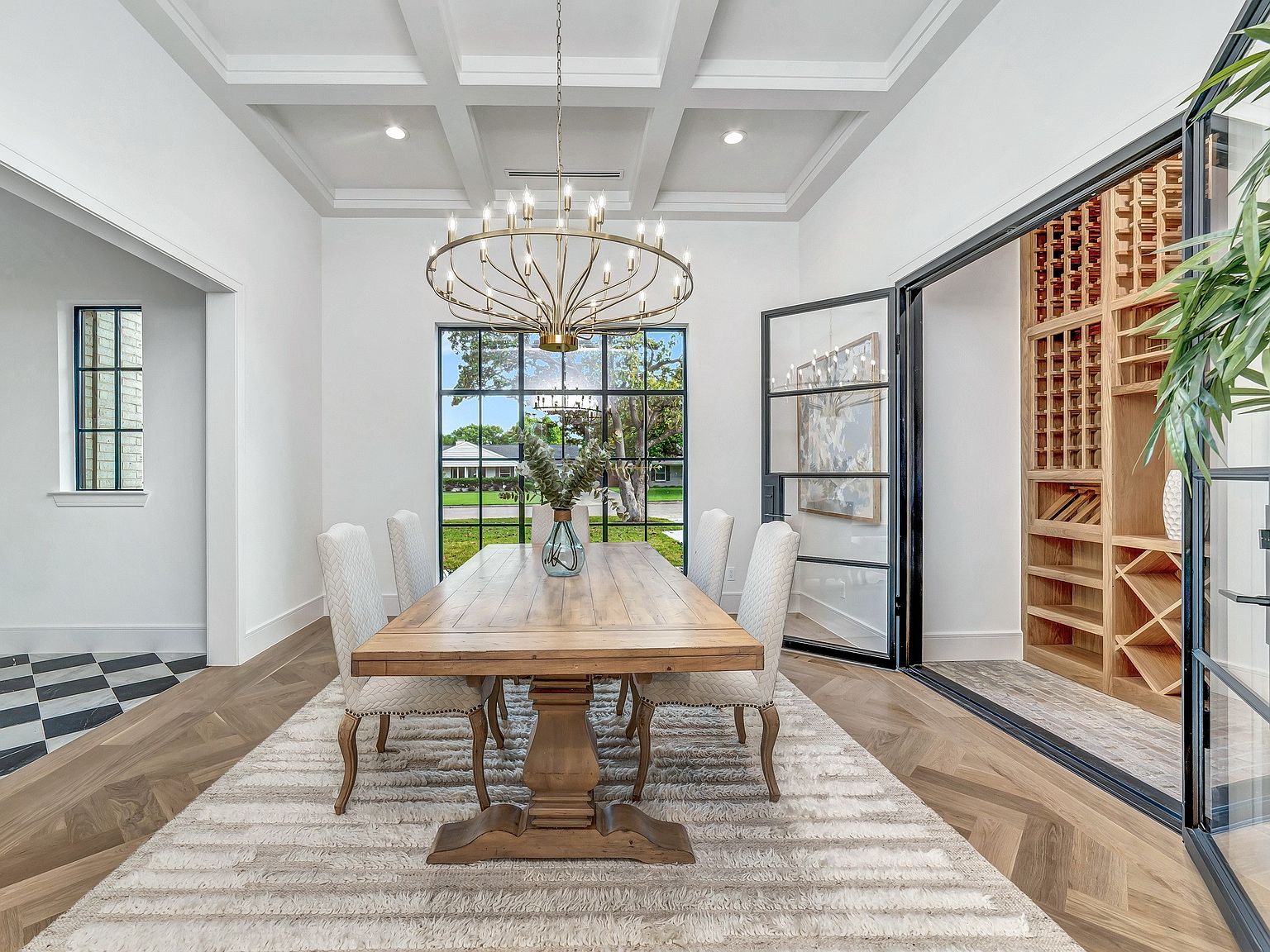 This is a bright and elegant dining room featuring a large wooden table with seating for six, illuminated by a statement chandelier. The room has white walls, a coffered ceiling, and a large window offering natural light and a view of the outdoors. A wine cellar is visible through a glass door, adding a luxurious touch.