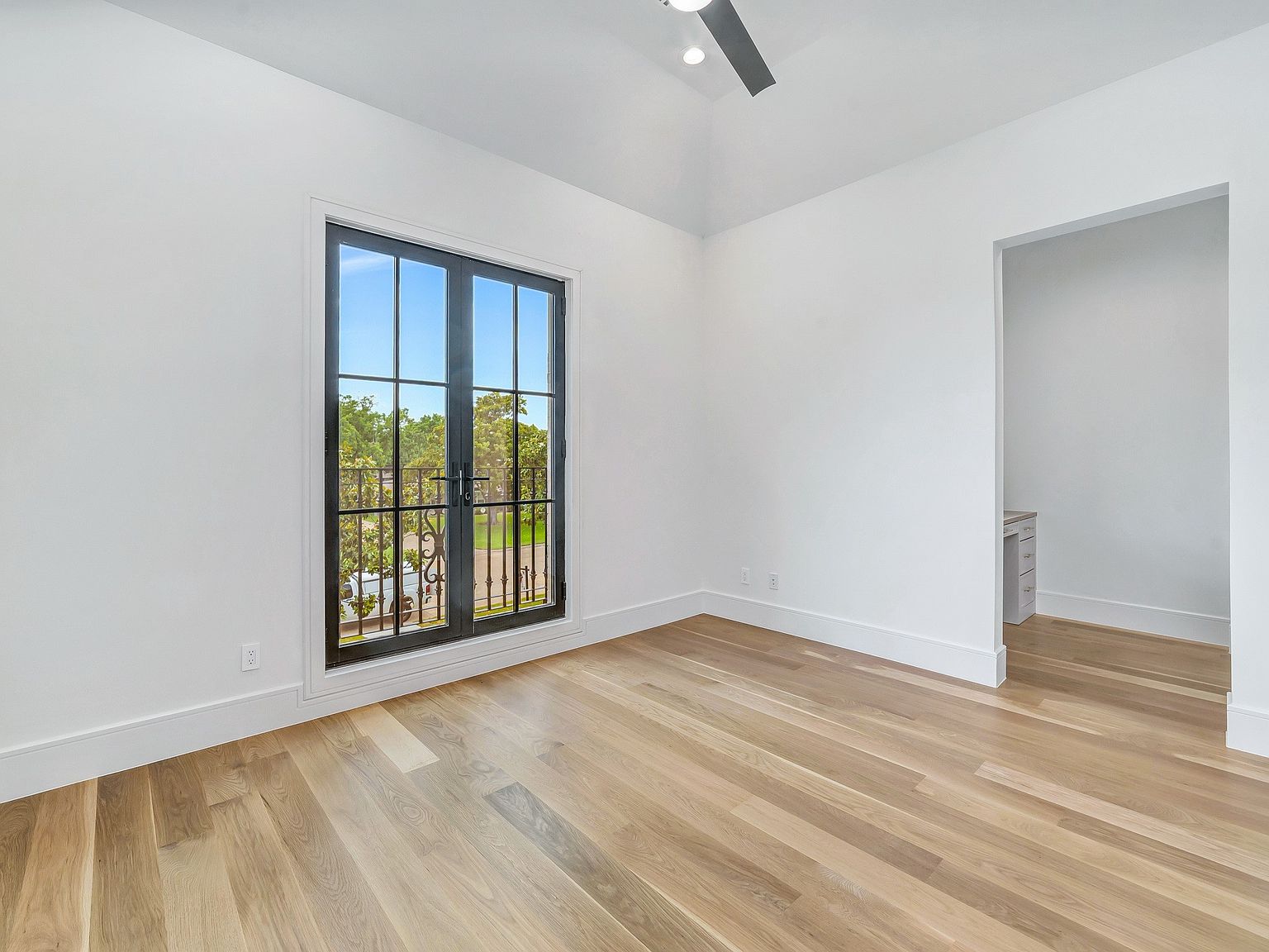 This is a bright and airy bedroom featuring light hardwood floors, white walls, and a black framed double door leading to a balcony. The room is minimalist in style, with clean lines and ample natural light, creating a serene and inviting atmosphere. The perspective is from the corner of the room, showcasing the door and the adjacent wall.