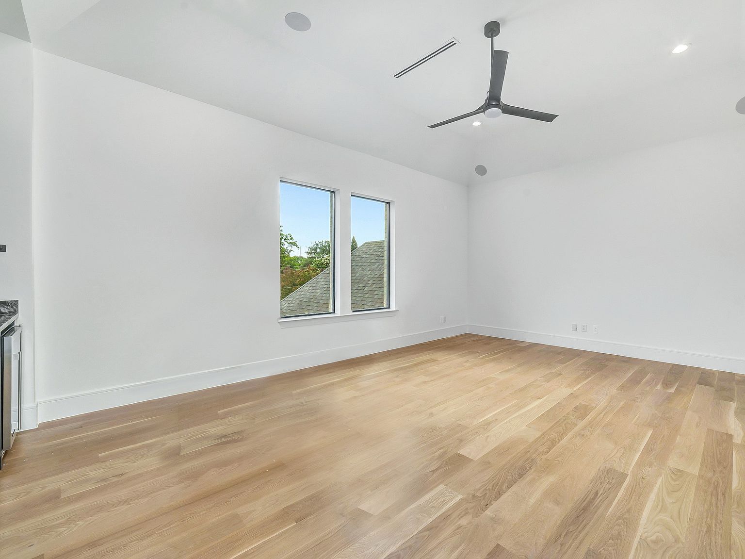 This is a bright and spacious living room featuring light hardwood floors and white walls, creating a modern and airy feel. Natural light floods the room through two windows, and a ceiling fan adds to the comfort. The room is empty, showcasing its size and potential for customization.