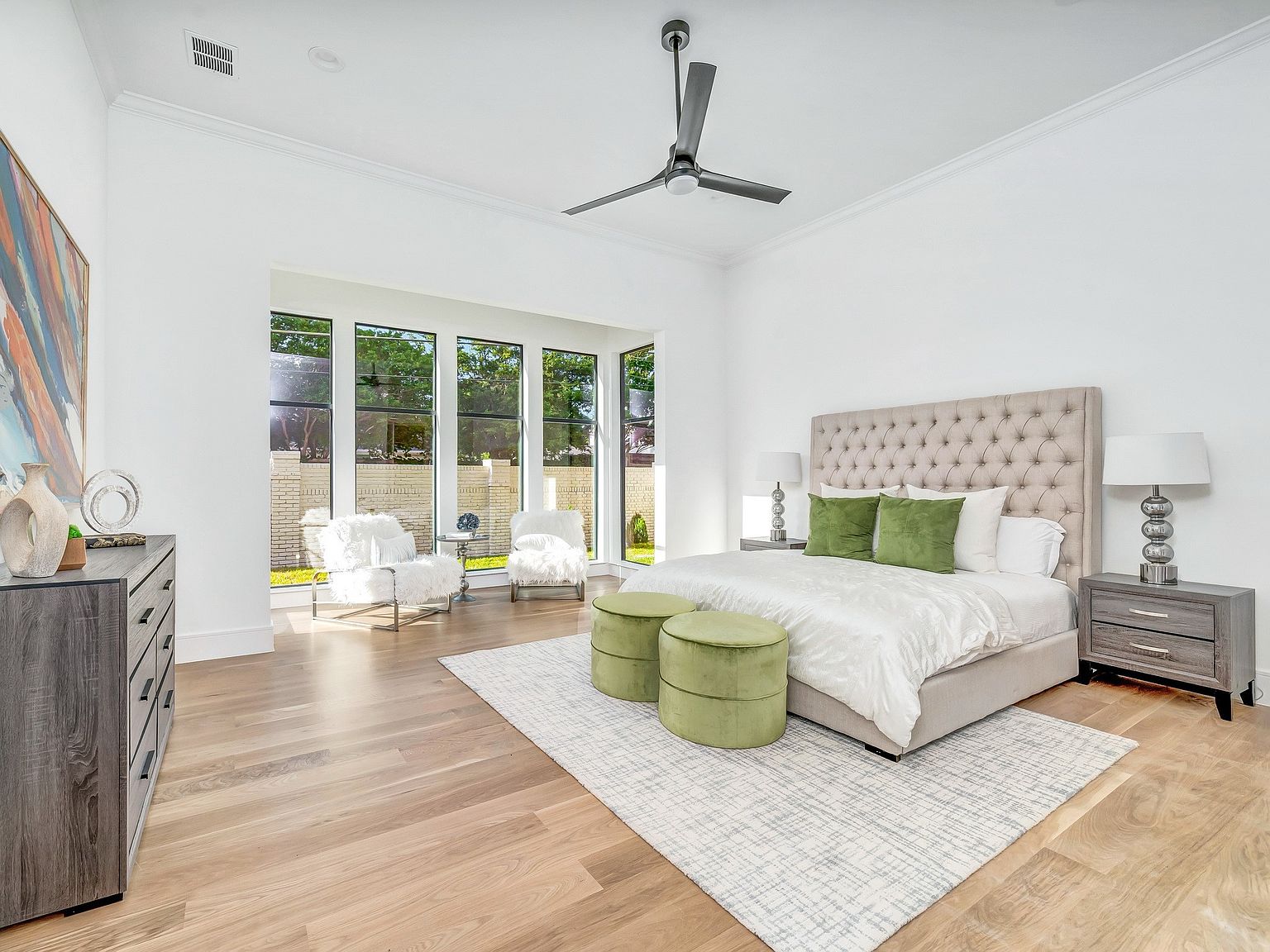 This is a bright and spacious primary bedroom featuring a large tufted headboard, neutral color palette, and hardwood floors. The room is well-lit with natural light from a large window and complemented by stylish bedside lamps. Green ottomans and pillows add a pop of color, while a patterned rug anchors the bed, creating a serene and luxurious atmosphere.