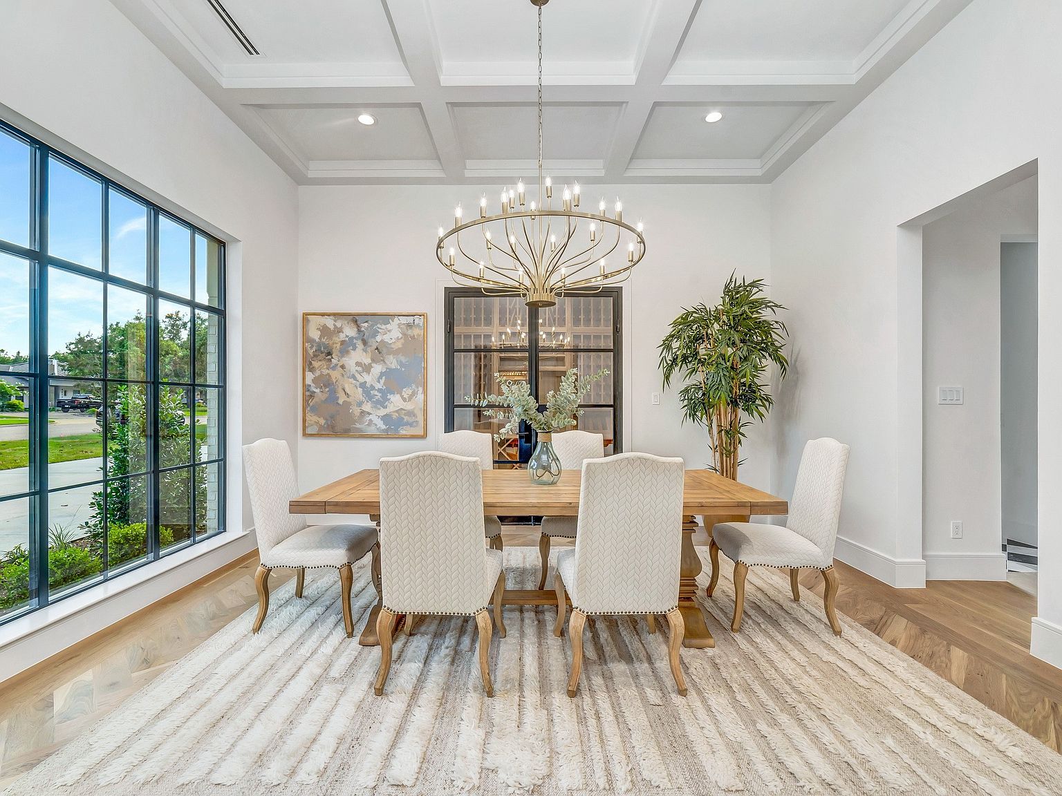 This is a bright and elegant dining room featuring a large wooden table surrounded by six upholstered chairs. A striking chandelier hangs above the table, and a large window provides natural light and a view of the outdoors. The room is decorated in neutral tones with a textured rug, creating a sophisticated and inviting atmosphere.