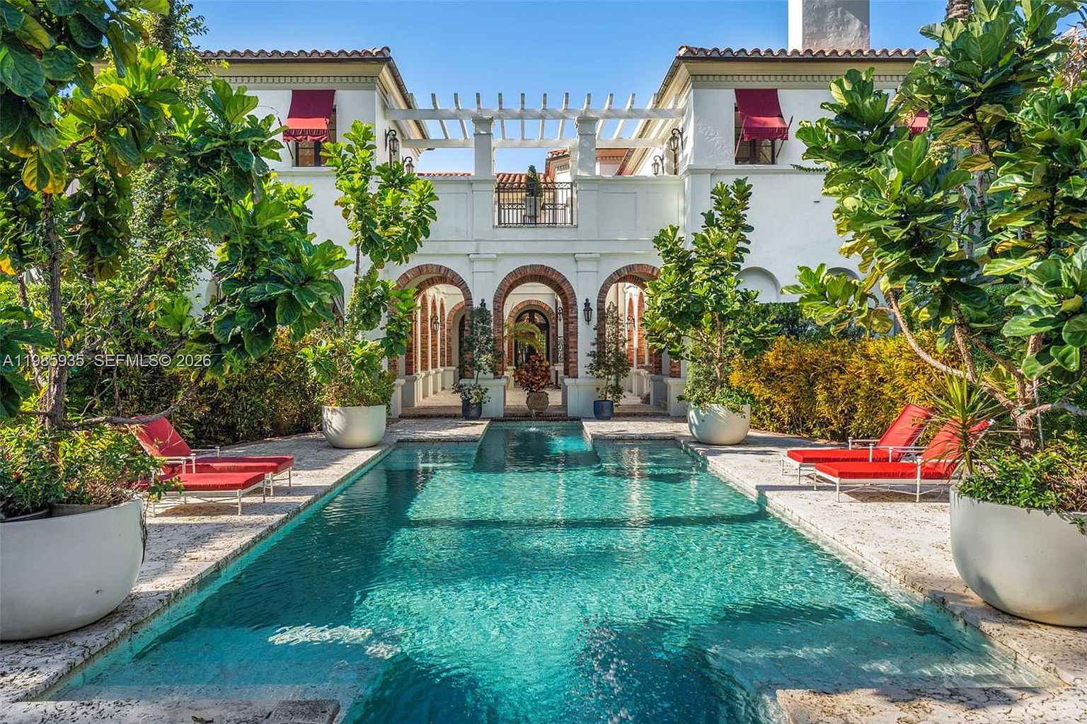 This Mediterranean-style estate features a stunning rectangular swimming pool as the centerpiece of a lush, private courtyard. The home showcases elegant architectural details, including arched loggias with brick accents, a second-story balcony, and a pergola-covered terrace. Large potted plants and vibrant greenery frame the pool area, creating a serene and luxurious outdoor living space.