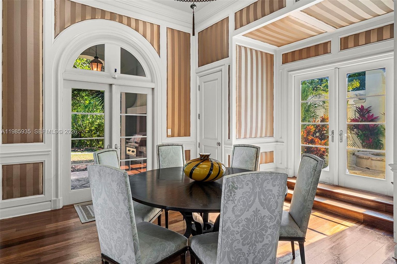 This elegant dining room features a dark circular table surrounded by upholstered chairs with a subtle damask pattern. The room is defined by its sophisticated wall treatment, which alternates between striped wallpaper panels and white wainscoting, complemented by a matching striped ceiling. Large glass doors on two sides provide abundant natural light and views of the lush outdoor greenery, creating a bright and airy atmosphere.
