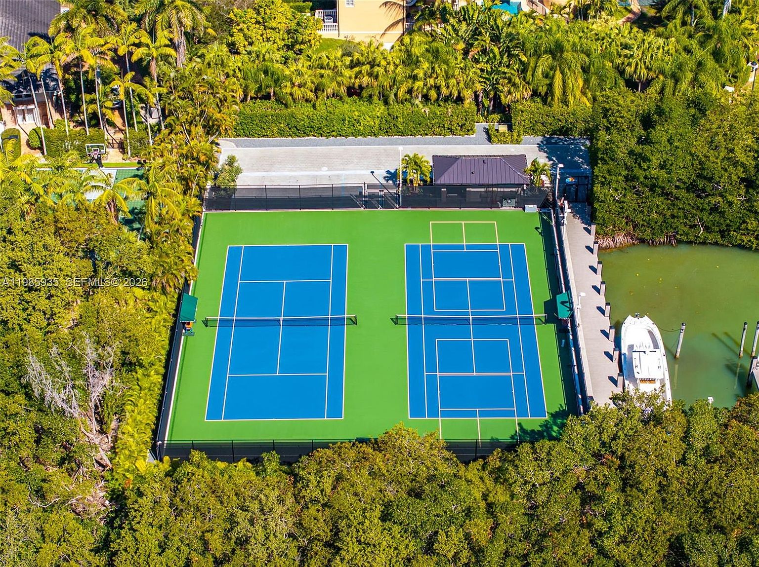 This high-angle aerial view showcases a vibrant green and blue tennis court nestled within a lush, tree-lined property. Adjacent to the court is a paved area with a covered pavilion and a private dock extending into a calm, green-toned waterway where a small boat is moored. The scene captures a serene, recreational outdoor space surrounded by dense tropical foliage.