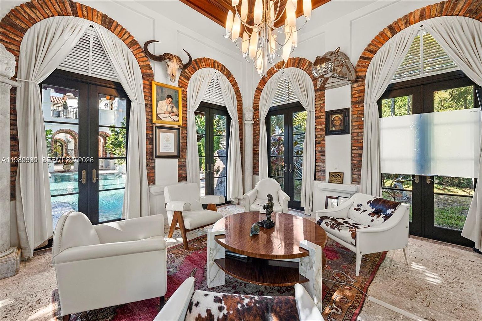 This elegant living room features a sophisticated blend of classic and eclectic design, highlighted by four arched doorways framed in exposed brick that lead to a pool area. The space is furnished with a mix of white leather armchairs and cowhide-patterned seating arranged around a circular wooden coffee table. A grand chandelier hangs from the ceiling, casting light over the patterned area rug and stone flooring, creating a luxurious and inviting atmosphere.