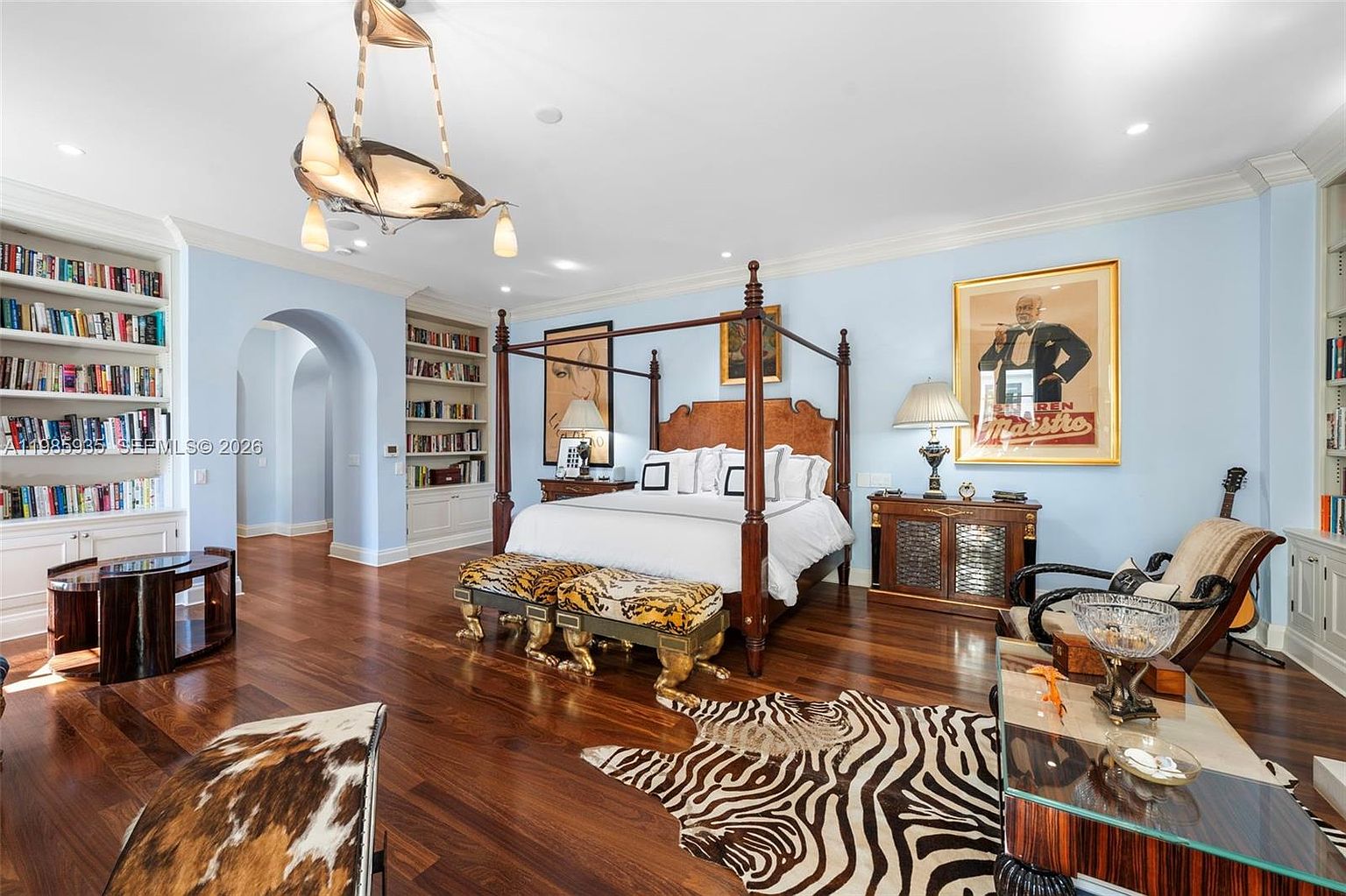 This spacious primary bedroom features a grand four-poster bed as the centerpiece, complemented by a zebra-print area rug and two matching animal-print benches at the foot of the bed. The room is accented by built-in bookshelves, a unique sculptural chandelier, and a framed vintage-style poster on the wall. The warm wood flooring and light blue walls create a sophisticated and inviting atmosphere.