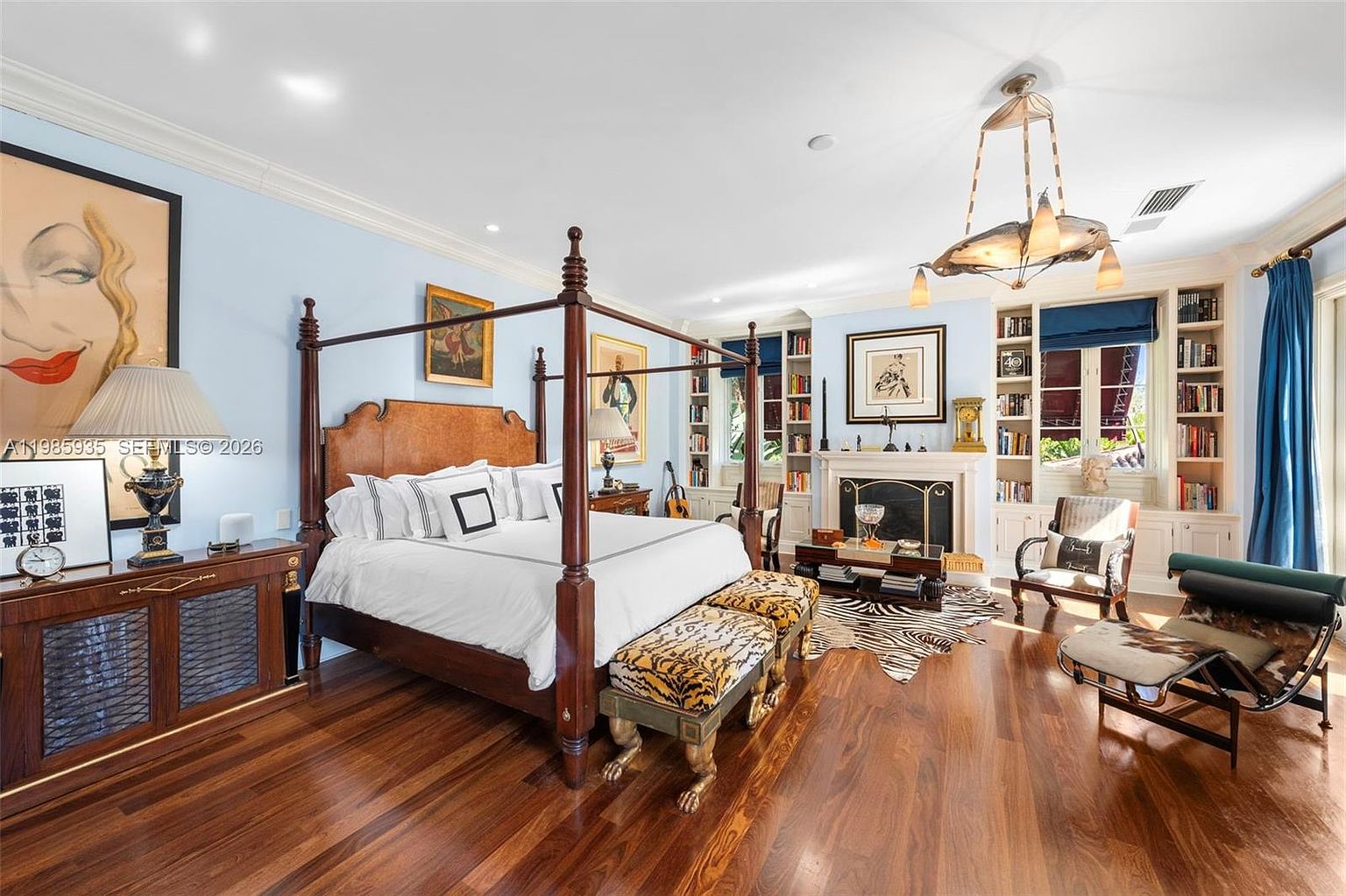 This spacious primary bedroom features rich, dark wood flooring and a prominent four-poster bed as the central focal point. The room is accented with eclectic decor, including animal-print benches, a zebra-patterned area rug, and a fireplace with a traditional mantel. Large windows with blue drapery and built-in shelving units flank the fireplace, creating a sophisticated and well-appointed living space.