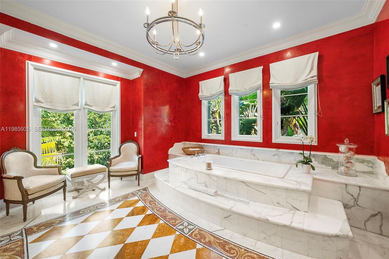 This luxurious primary bathroom features a prominent built-in soaking tub encased in white stone with intricate veining, set against a striking red wall. The room is accented by a classic chandelier, a patterned tile floor, and two elegant armchairs placed near large windows that provide ample natural light. The overall aesthetic is opulent and traditional, creating a sophisticated spa-like atmosphere.