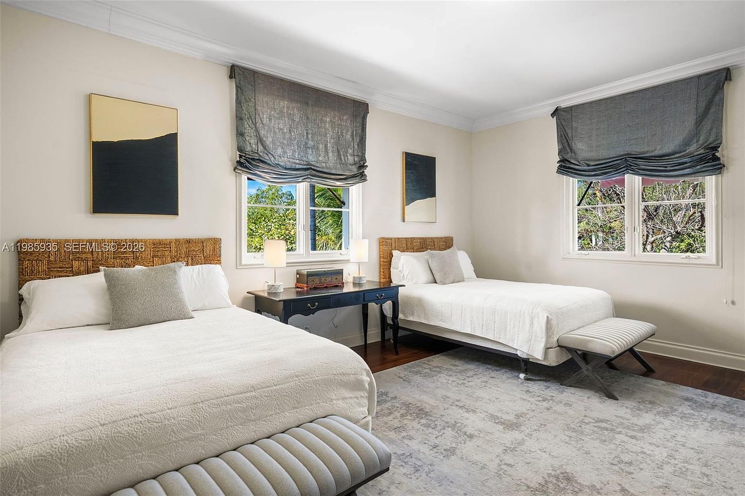 This bright and airy guest bedroom features two beds with woven headboards, crisp white bedding, and matching grey accent pillows. A dark wood console table sits between the beds, topped with two lamps and a decorative box, while large windows with grey Roman shades allow for ample natural light. The room is finished with a neutral patterned area rug and two upholstered benches at the foot of each bed, creating a cohesive and inviting space.