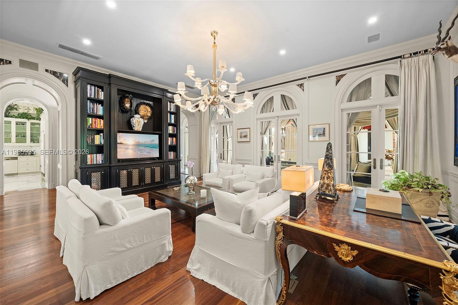 This elegant living room features dark wood flooring and a prominent black built-in shelving unit that houses a television and decorative items. The space is furnished with plush white seating arranged around a dark wood coffee table, all centered beneath a large, multi-armed chandelier. Tall arched glass doors provide natural light and views into an adjacent space, creating a sophisticated and airy atmosphere.