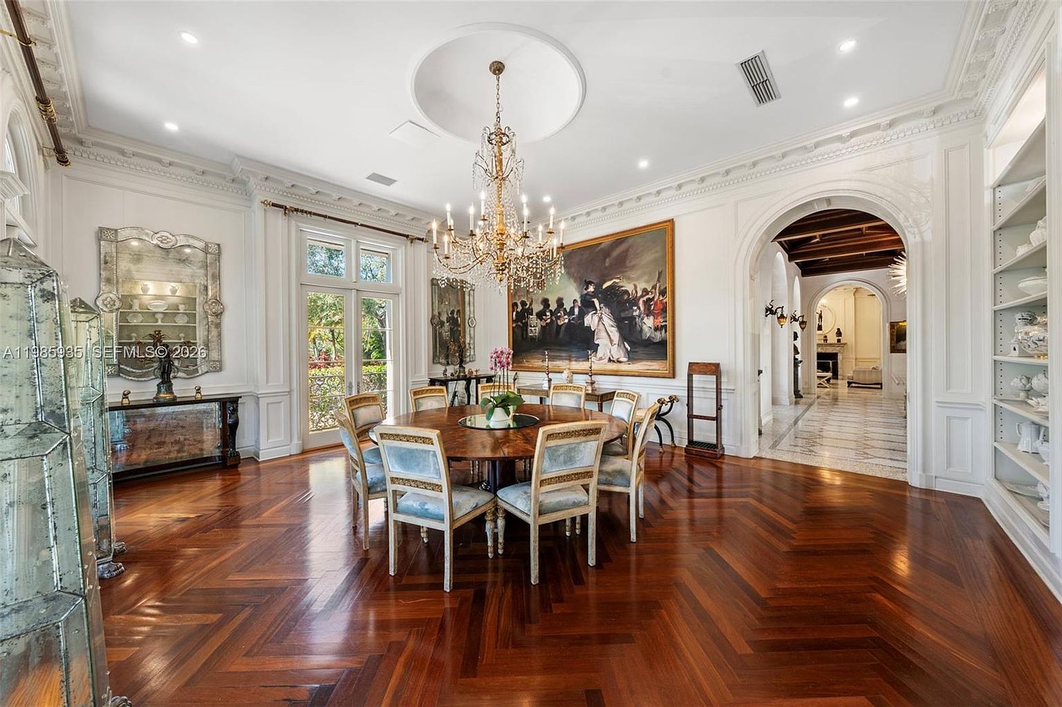 This elegant formal dining room features a large circular wooden table surrounded by upholstered chairs, centered beneath a grand crystal chandelier. The space is defined by rich, dark herringbone-patterned wood flooring, intricate crown molding, and a large, ornate painting on the main wall. An arched doorway leads to a bright, marble-floored hallway, while a tall, built-in shelving unit and a decorative mirror add to the sophisticated, classic aesthetic.