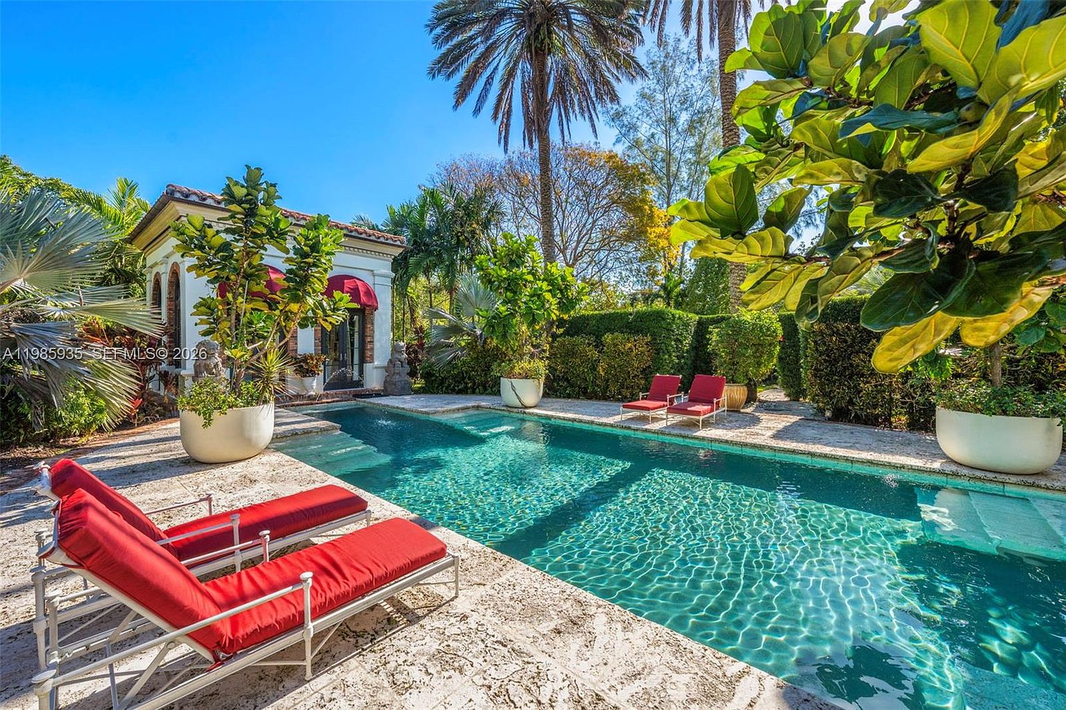 This serene outdoor space features a rectangular swimming pool surrounded by a light-toned stone patio. Four red lounge chairs are arranged around the pool, which is framed by lush tropical landscaping, including tall palm trees and dense hedges. A small, elegant pool house with red awnings sits at the far end, creating a private and luxurious resort-like atmosphere.