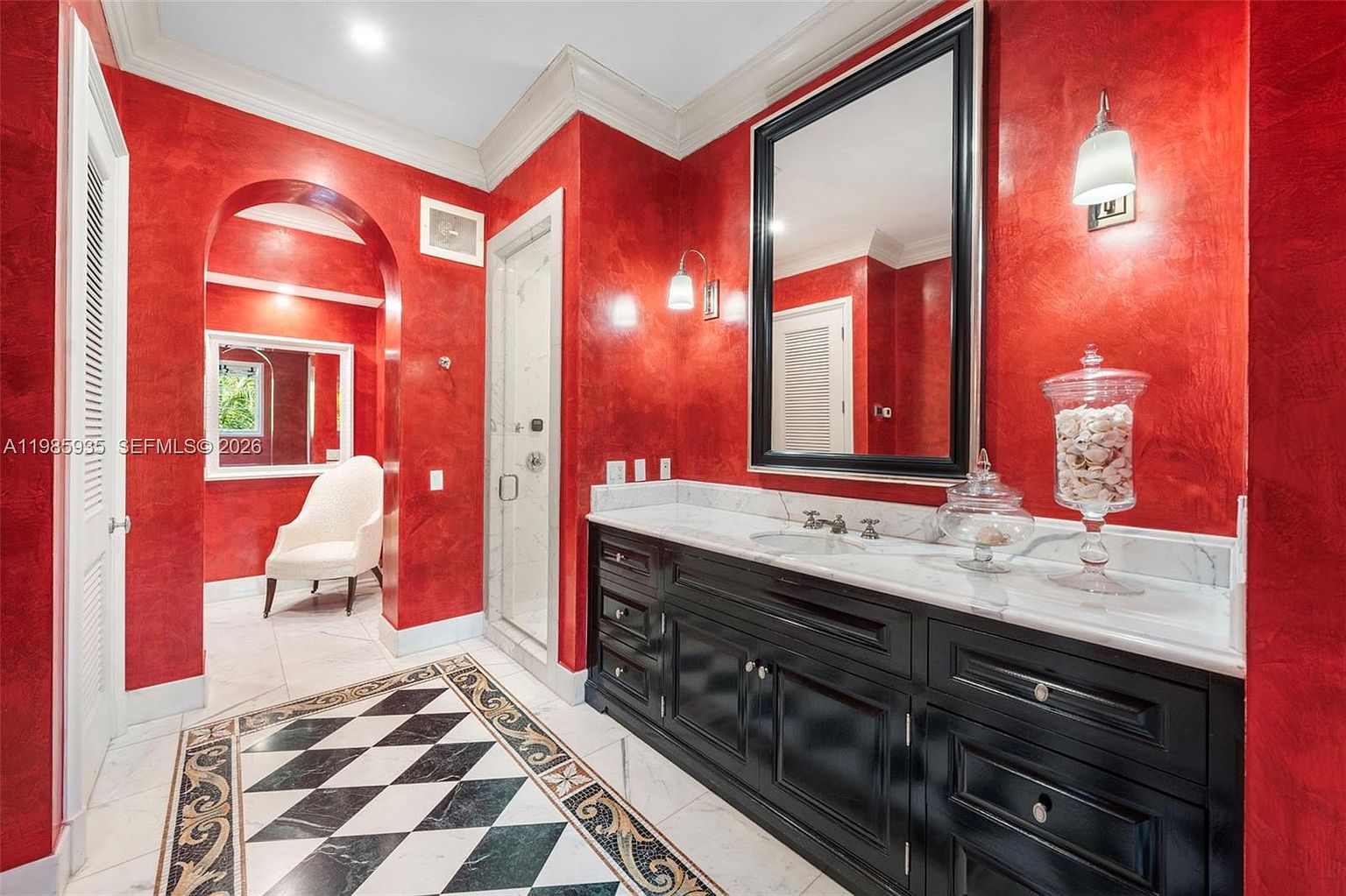 This primary bathroom features bold, textured red walls and a striking black vanity topped with a light-colored stone counter. The floor is adorned with a decorative black and white diamond-patterned tile inlay, while an arched doorway leads to a secondary space containing a white armchair. The perspective is a wide-angle shot from the doorway, capturing the contrast between the dark cabinetry and the vibrant wall color.