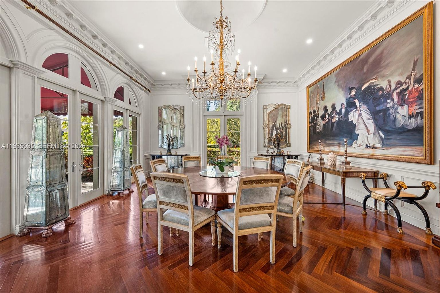 This elegant dining room features a large circular wooden table surrounded by upholstered chairs, set beneath a grand crystal chandelier. The space is defined by rich, dark wood flooring laid in a herringbone pattern and tall, arched glass doors that allow for abundant natural light. A massive, ornate painting hangs on the wall, complemented by classic architectural molding and decorative mirrored accents, creating a sophisticated and formal atmosphere.