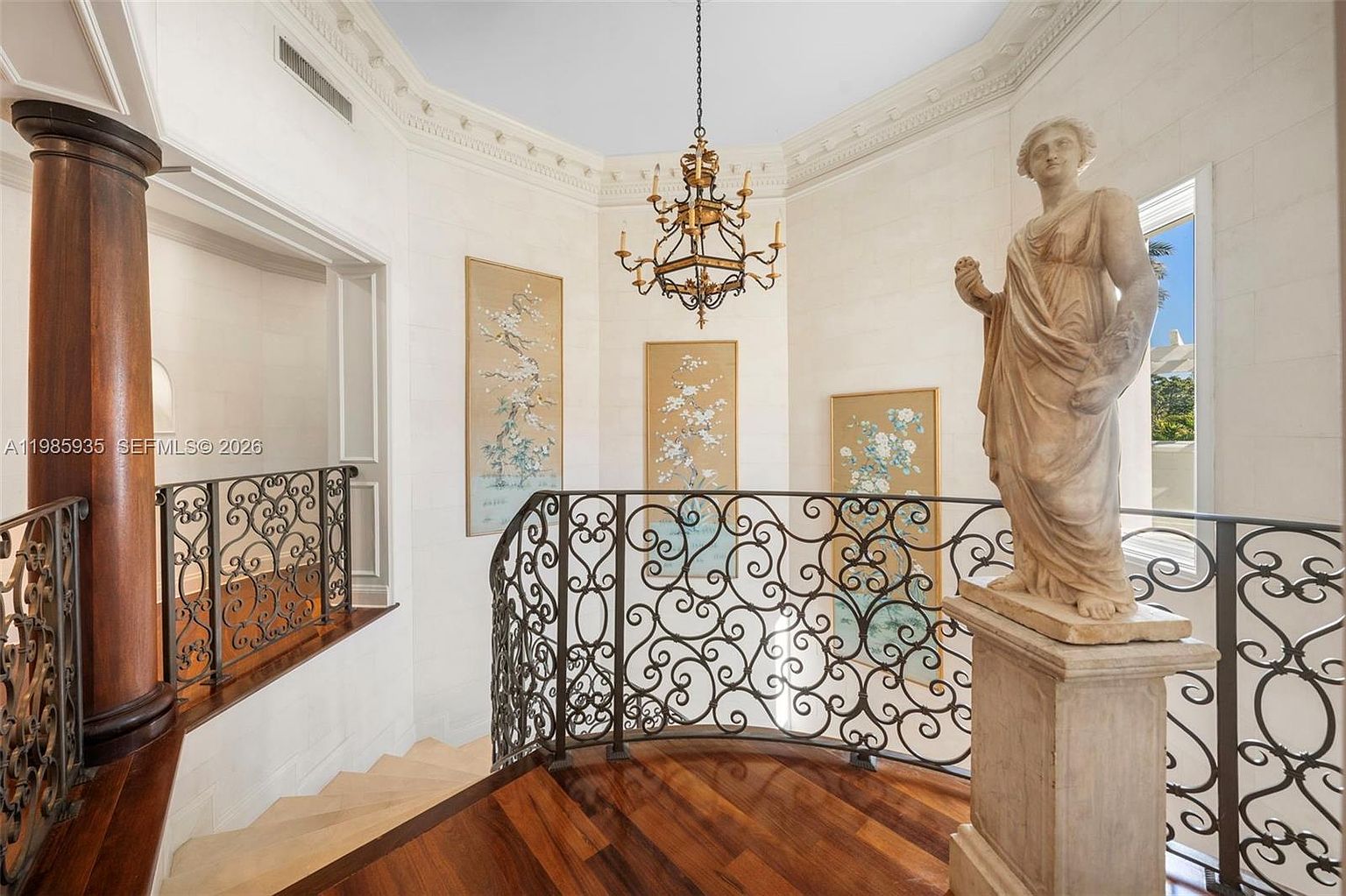 This elegant staircase landing features a curved wrought-iron railing and rich wood flooring, creating a sophisticated architectural focal point. A classical-style statue stands prominently on a pedestal, complemented by three framed botanical art pieces mounted on the light-toned walls. An ornate gold chandelier hangs from the ceiling, casting a warm glow over the space, which is framed by a large, dark wood column.
